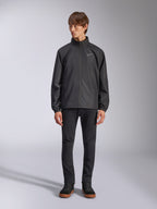 Alpinestars Overhaul Racer Jacke, schwarze Regenjacke für Herren, Ganzkörperansicht an einem Model kombiniert mit schwarzer Hose und lässigen Sneakern, mit kleinem weißen Logo auf der Brust, wasserabweisendem Material und lässigem, vom Motorsport inspiriertem Design