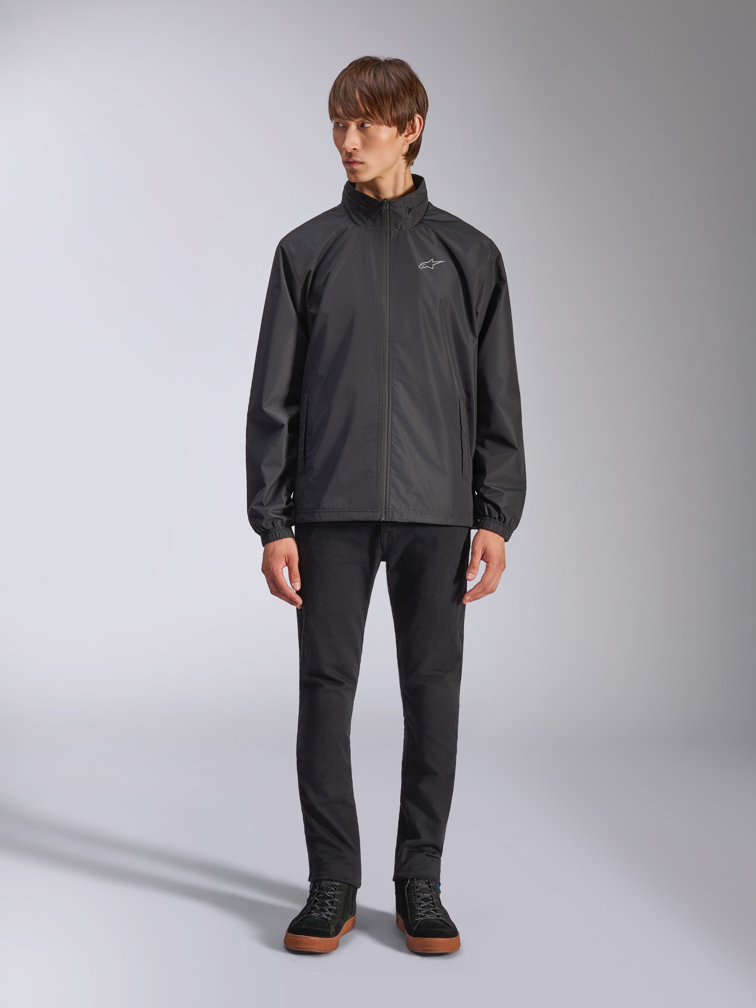 Alpinestars Overhaul Racer Jacke, schwarze Regenjacke für Herren, Ganzkörperansicht an einem Model kombiniert mit schwarzer Hose und lässigen Sneakern, mit kleinem weißen Logo auf der Brust, wasserabweisendem Material und lässigem, vom Motorsport inspiriertem Design
