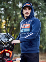Alpinestars Harken Hoodie, Navyblau, Fleece-Kapuzenpullover mit Brustgrafik mit Farbverlauf und weißem Alpinestars-Schriftzug am Ärmel, getragen von einem Mann, der einen Motorradhelm hält, lässige, vom Motorsport inspirierte Streetwear