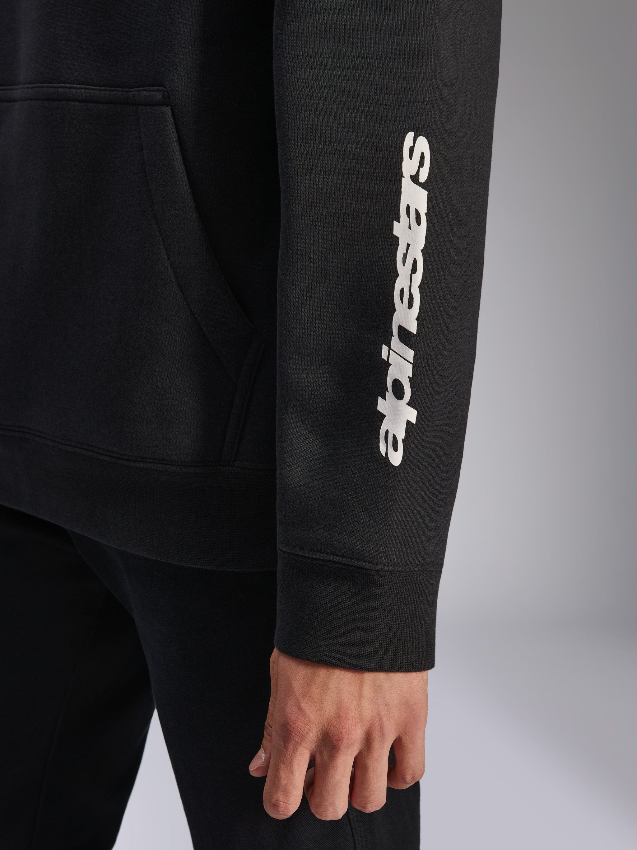 Alpinestars Harken Hoodie, Schwarz, Nahaufnahme des Ärmels mit vertikalem weißem Wortmarken-Logo, Rippbündchen und Kängurutaschen-Detail, lässiges Sportbekleidungs-Fleece