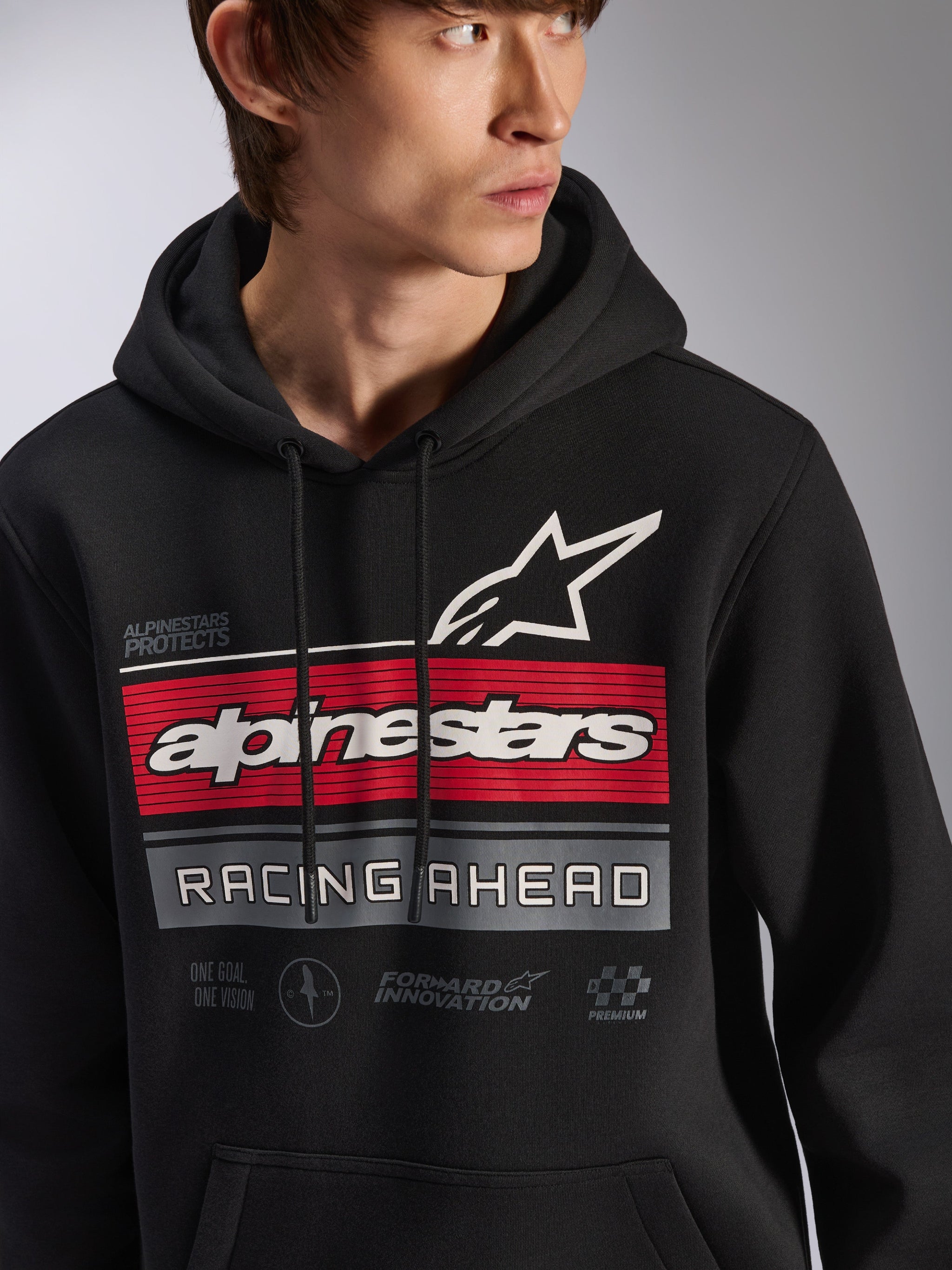 Alpinestars Harken Hoodie, schwarzer Fleece-Kapuzenpullover, getragen von einem Model, mit einer großen Grafik auf der Brust mit roten und grauen Streifen, weißem Alpinestars-Schriftzug und Stern-Logo, „Racing Ahead“-Schriftzug, moderne motorsport-inspirierte Casual-Streetwear.