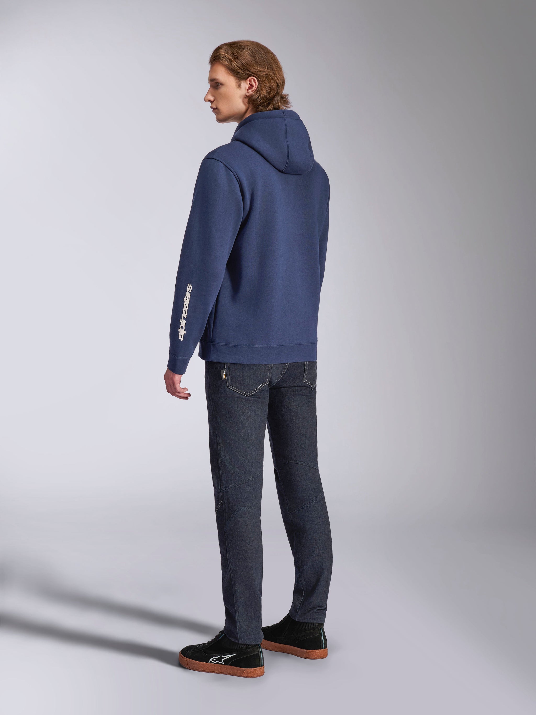 Alpinestars Harken Hoodie in Navy, Rückansicht eines männlichen Models, lässiger Fleece-Kapuzenpullover mit weißem Wortmarken-Logo am Ärmel, kombiniert mit dunkler Denim-Jeans und schwarzen Sneakern, moderne Motorsport-inspirierte Streetwear