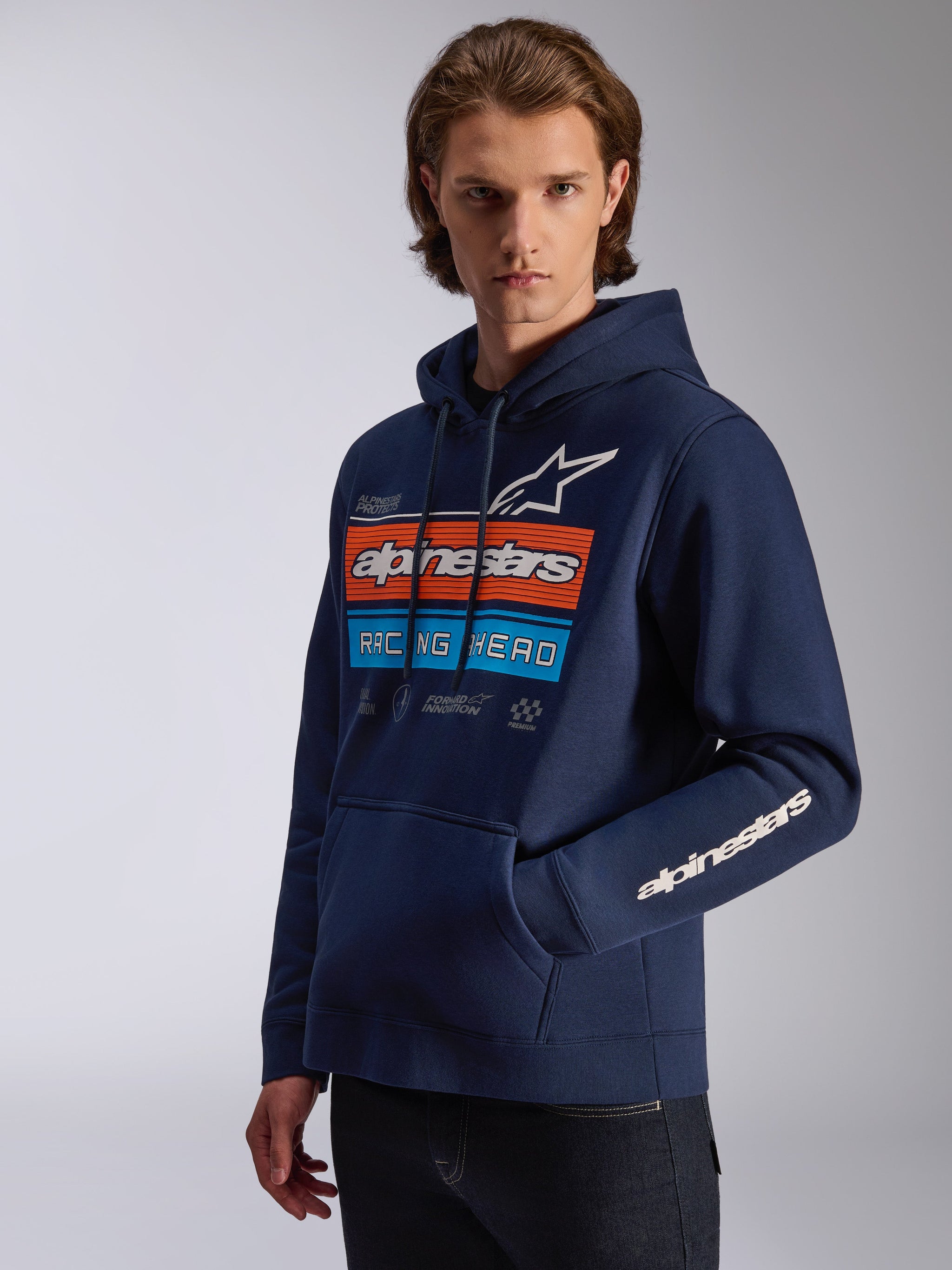 Alpinestars Harken Hoodie, Marineblau, von einem Modell getragen, mit einer markanten Brustgrafik im orange-blauen 'Racing Ahead'-Design, Alpinestars-Wortmarke auf dem Ärmel, Kängurutasche und Kapuze mit Kordelzug