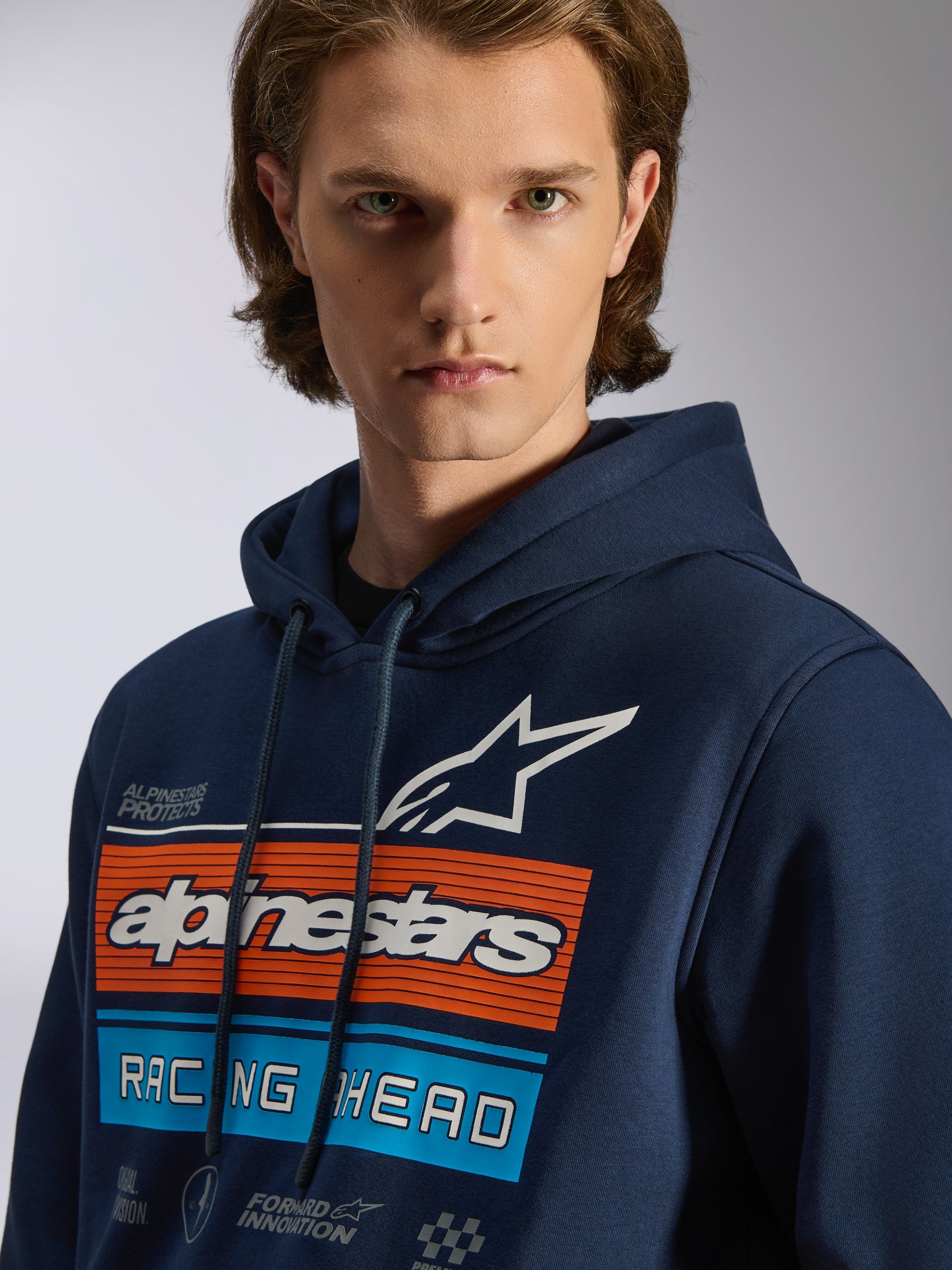 Alpinestars Harken Hoodie, dunkelblauer Kapuzenpullover, Brustgrafik mit orangefarbenen und hellblauen horizontalen Balken, weißes Stern-Logo, „Racing Ahead“-Schriftzug, lässige Motorsport-Streetwear