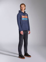 Alpinestars Harken Hoodie in Marineblau, getragen von einem männlichen Model mit dunkler Denim-Hose und schwarzen Sneakern, mit einer großen Brustgrafik mit orangefarbenen und hellblauen Akzenten, Alpinestars-Wortmarke und Kängurutasche.