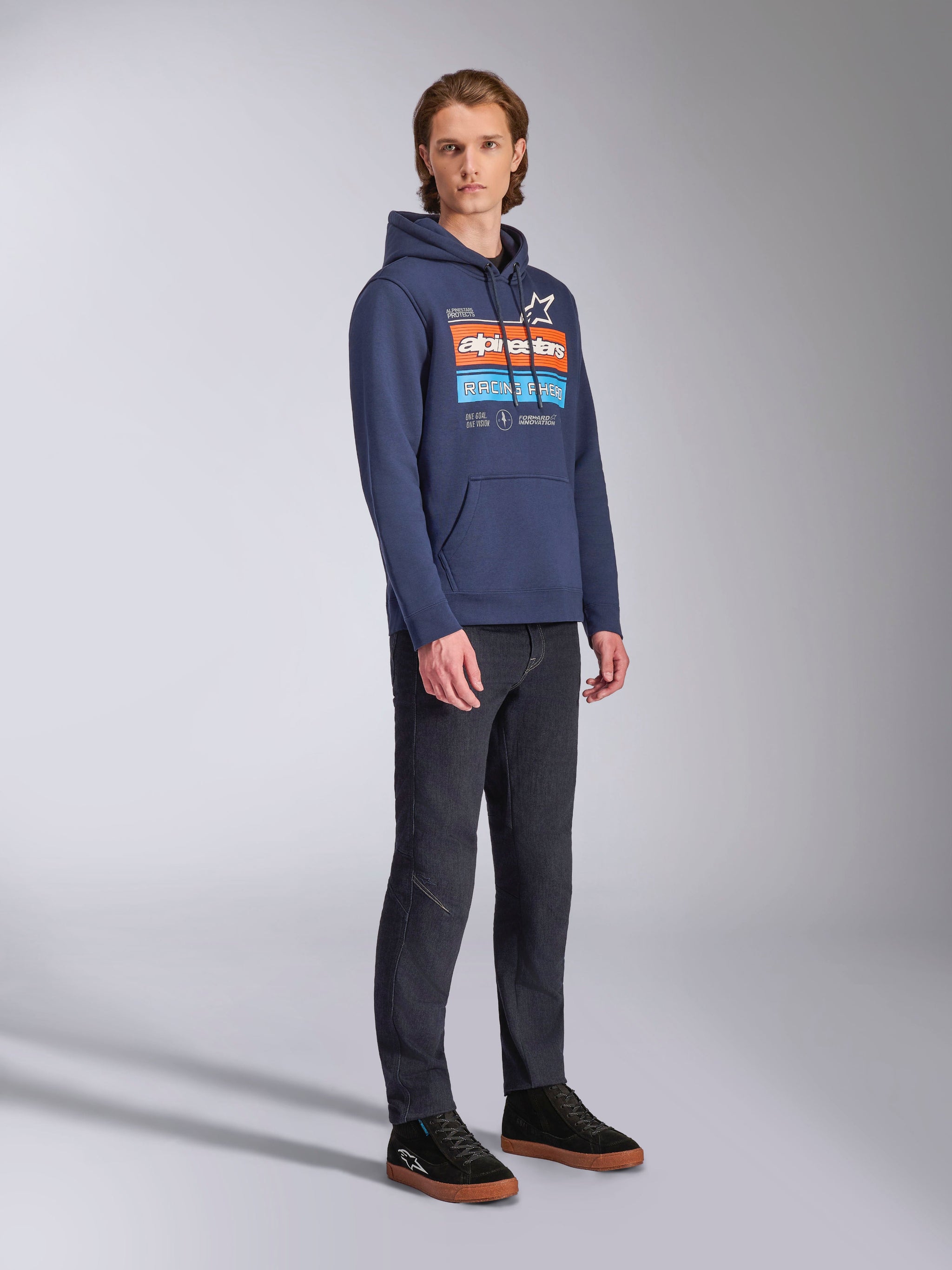 Alpinestars Harken Hoodie in Marineblau, getragen von einem männlichen Model mit dunkler Denim-Hose und schwarzen Sneakern, mit einer großen Brustgrafik mit orangefarbenen und hellblauen Akzenten, Alpinestars-Wortmarke und Kängurutasche.
