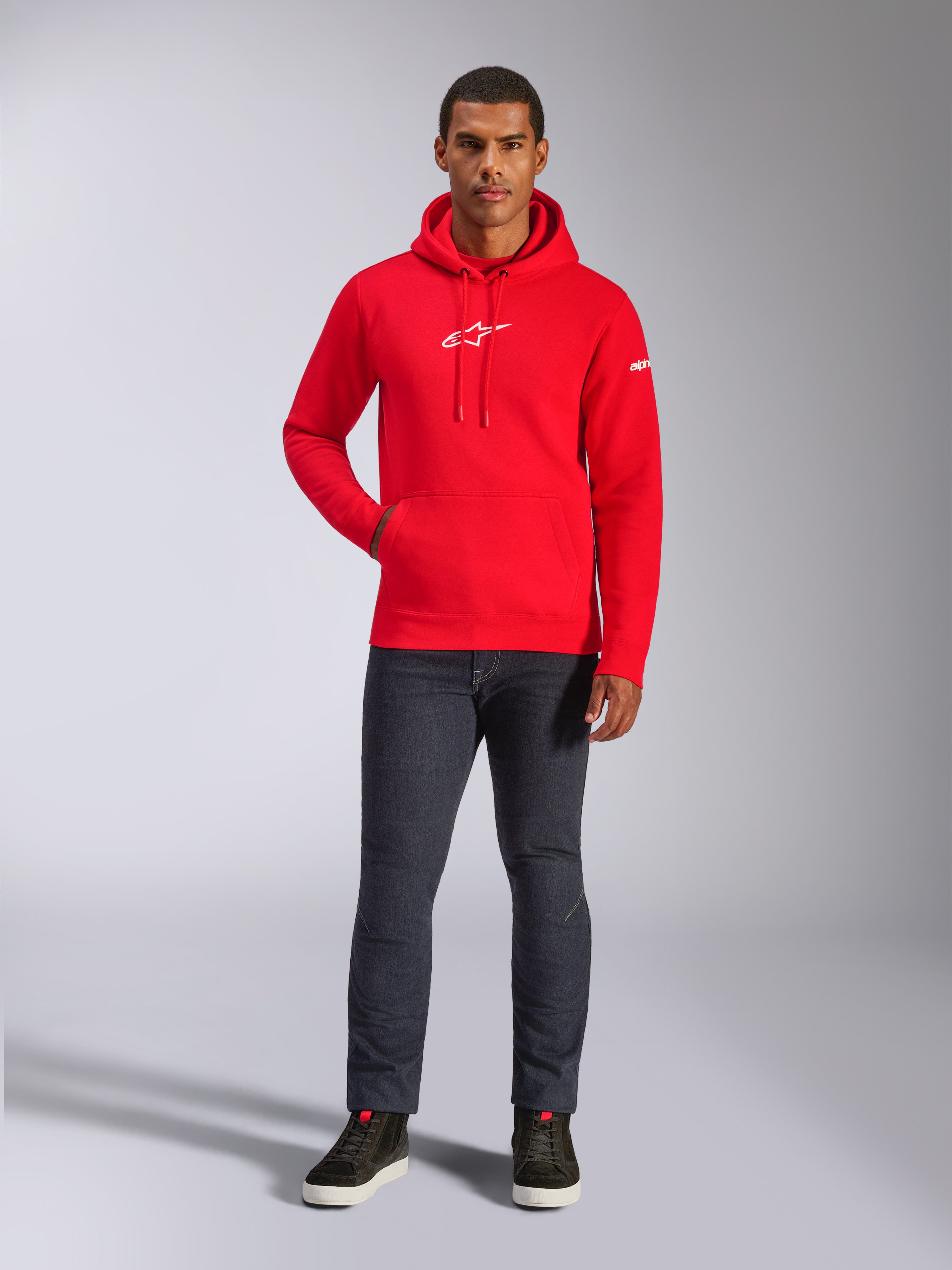 Männliches Model trägt den Alpinestars Frontal Hoodie in Rot, Pullover-Fleece-Hoodie mit weißem Astars-Logo auf der Brust, Kängurutasche, Kapuze mit Kordelzug, kombiniert mit dunkelgrauer Jeans und schwarzen High-Top-Sneakern, lässige Motorsport-Streetwear