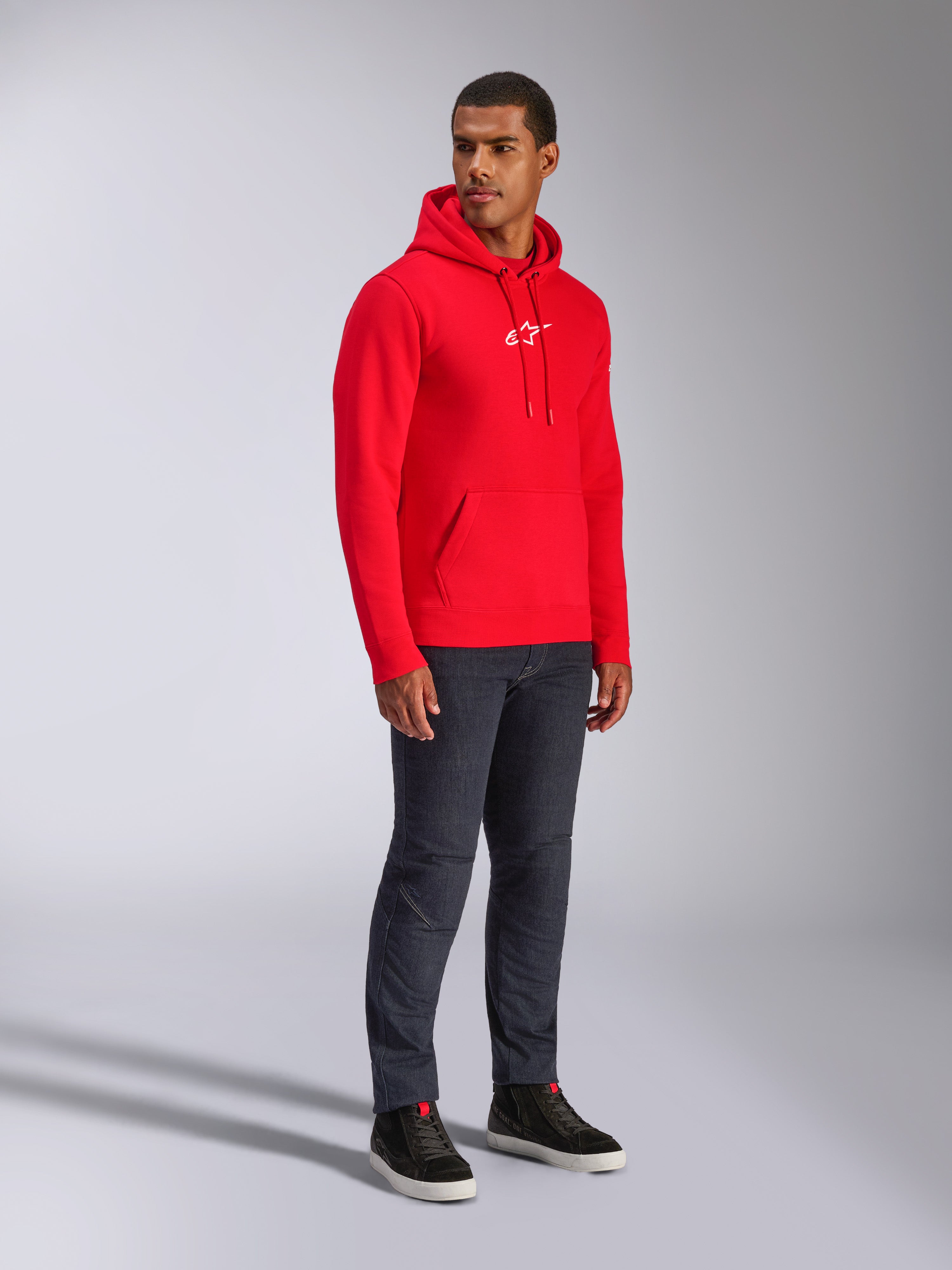 Alpinestars Frontal Hoodie, Rot, männliches Model trägt Fleece-Kapuzenpullover mit weißem A-Star-Brustlogo, Kängurutasche, Kapuze mit Kordelzug, kombiniert mit dunklen Denim-Jeans, lässige vom Motorsport inspirierte Streetwear