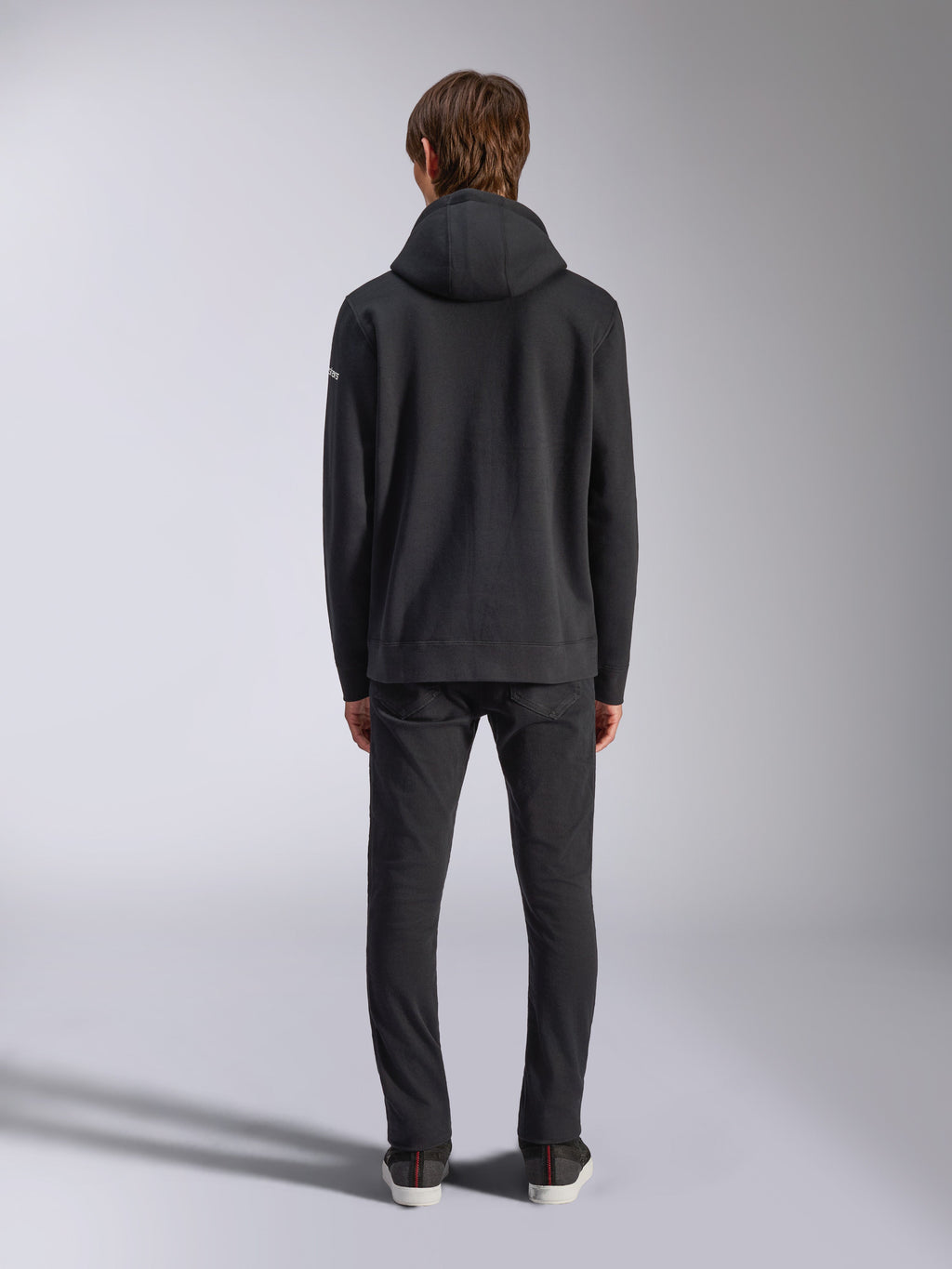 Alpinestars Frontal Hoodie, lässige Streetwear, Schwarz, Rückansicht eines Models im Fleece-Kapuzenpullover, mit Rippbündchen an Saum und Ärmeln, kleines weißes Wortmarken-Logo am Ärmel, kombiniert mit schwarzer Hose