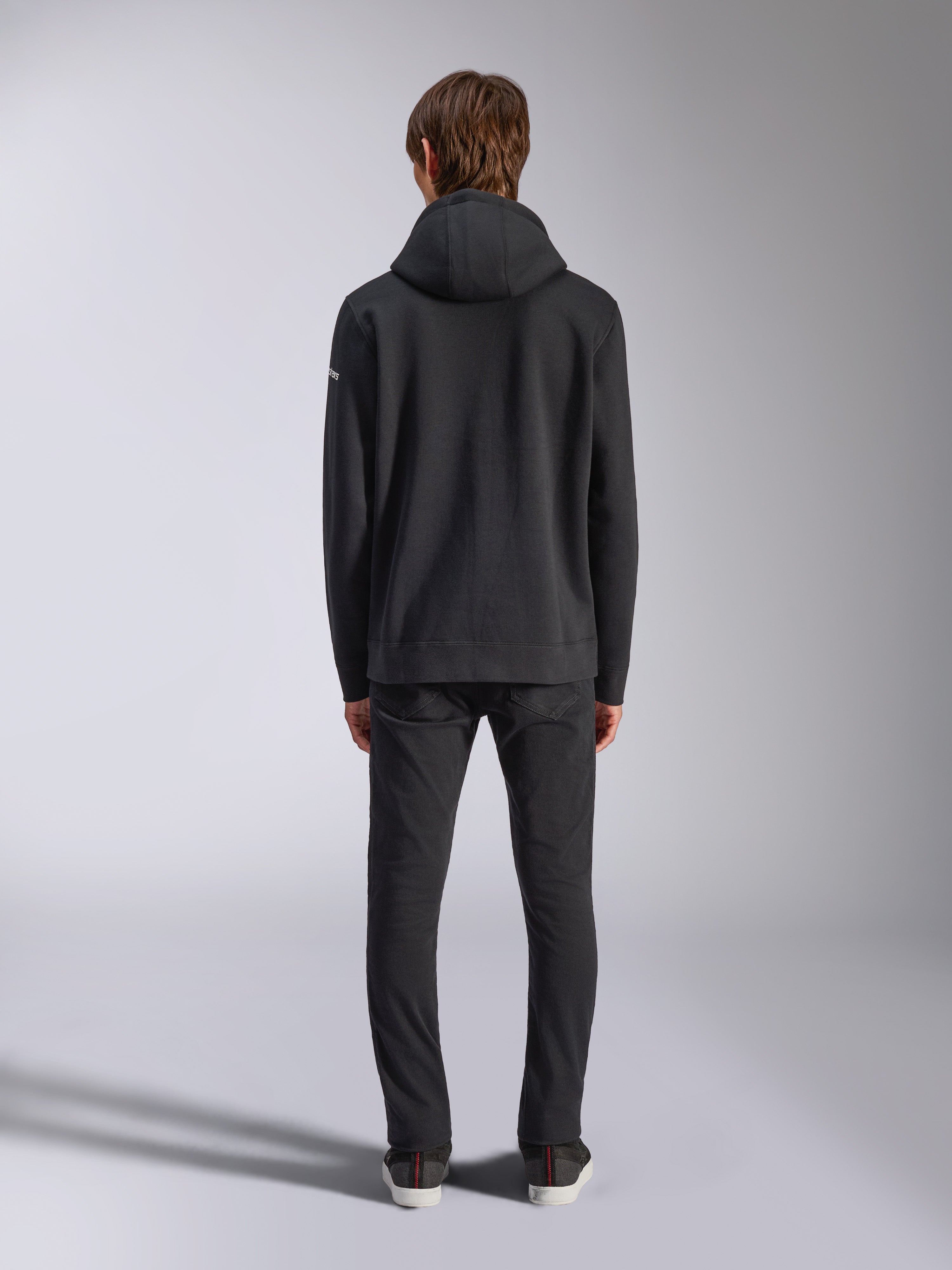 Alpinestars Frontal Hoodie, lässige Streetwear, Schwarz, Rückansicht eines Models im Fleece-Kapuzenpullover, mit Rippbündchen an Saum und Ärmeln, kleines weißes Wortmarken-Logo am Ärmel, kombiniert mit schwarzer Hose
