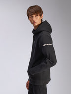 Alpinestars Frontal Hoodie, schwarzer Fleece-Pullover, Modell in Seitenansicht, weißer Alpinestars-Schriftzug auf dem Ärmel, Kängurutasche, lässige, vom Motorsport inspirierte Streetwear
