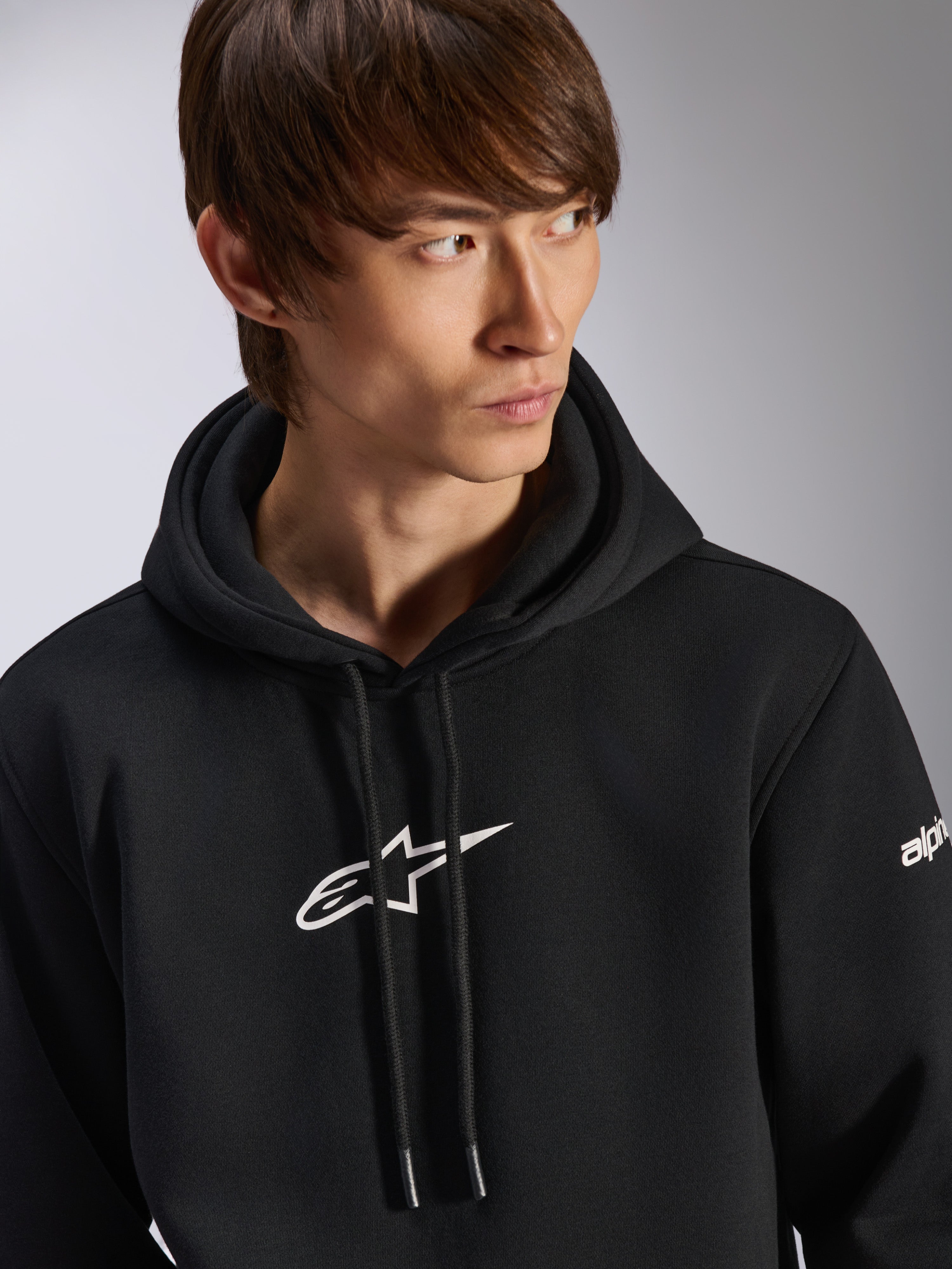 Alpinestars Frontal Hoodie, schwarzer Fleece-Kapuzenpullover, weißes Astar-Logo auf der Brust und Wortmarken-Branding am Ärmel, Kapuze mit Kordelzug, getragen von einem männlichen Model, lässige Motorsport-Streetwear