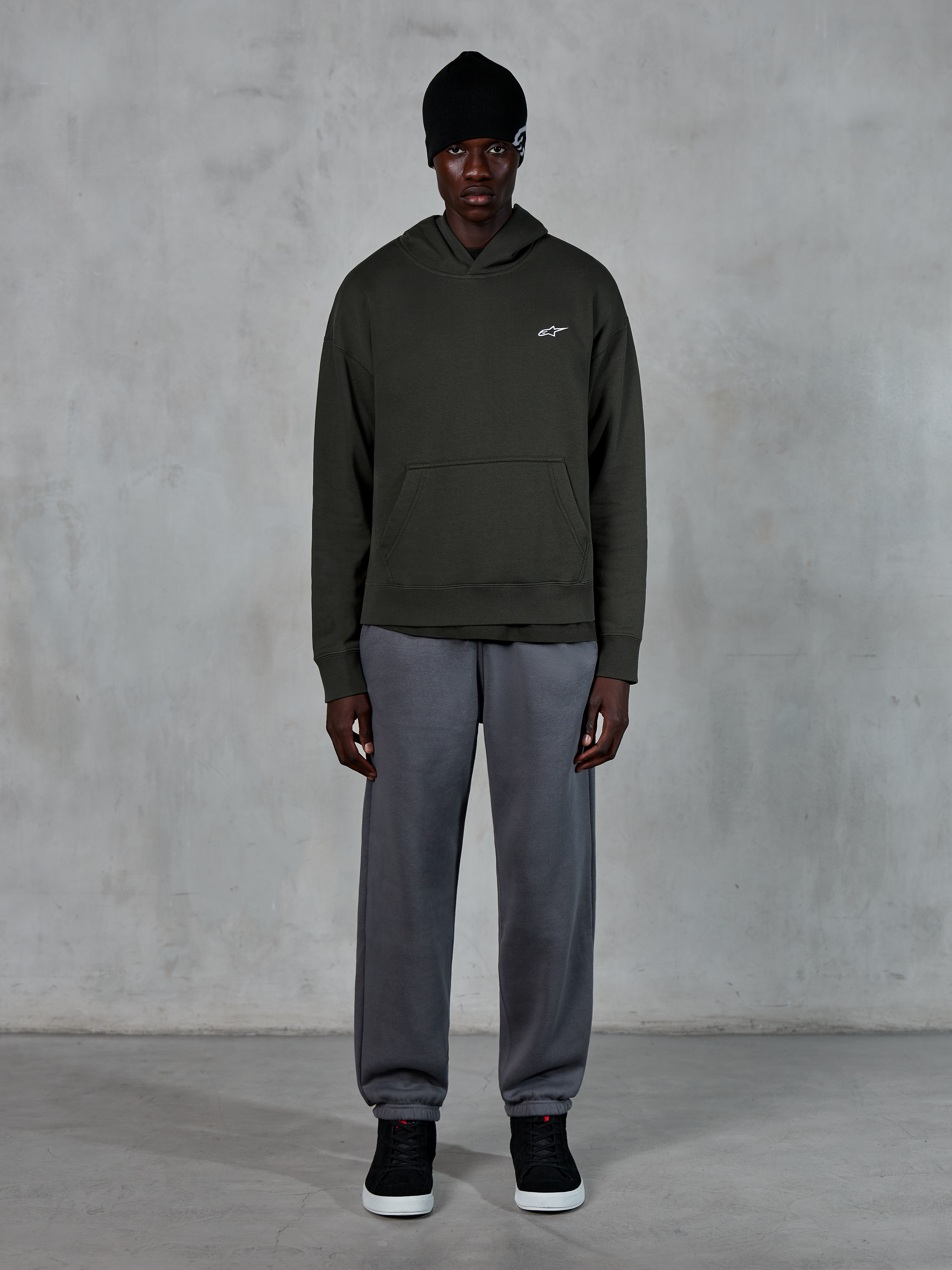 Model trägt Alpinestars Essentials Ageless Hoodie, lässige Streetwear, Off Black mit kleinem weißem Logo auf der Brust, kombiniert mit grauer Jogginghose, schwarzer Beanie und schwarzen Sneakern, minimalistisches, vom Motorsport inspiriertes Design