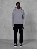 Alpinestars Essentials Ageless Hoodie, Silber, männliches Model in lässiger Streetwear, kleines schwarzes Alpinestars-Logo auf der Brust, Kängurutasche vorne, minimalistisches Design, kombiniert mit schwarzer Hose und Sneakern.