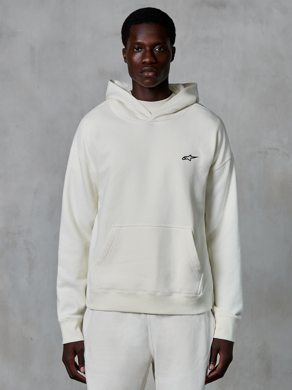 Alpinestars Essentials Ageless Hoodie, Off-White, getragen von einem männlichen Model, kleines schwarzes Logo auf der Brust, Kängurutasche vorne, lange Ärmel, lässige, vom Motorsport inspirierte Streetwear