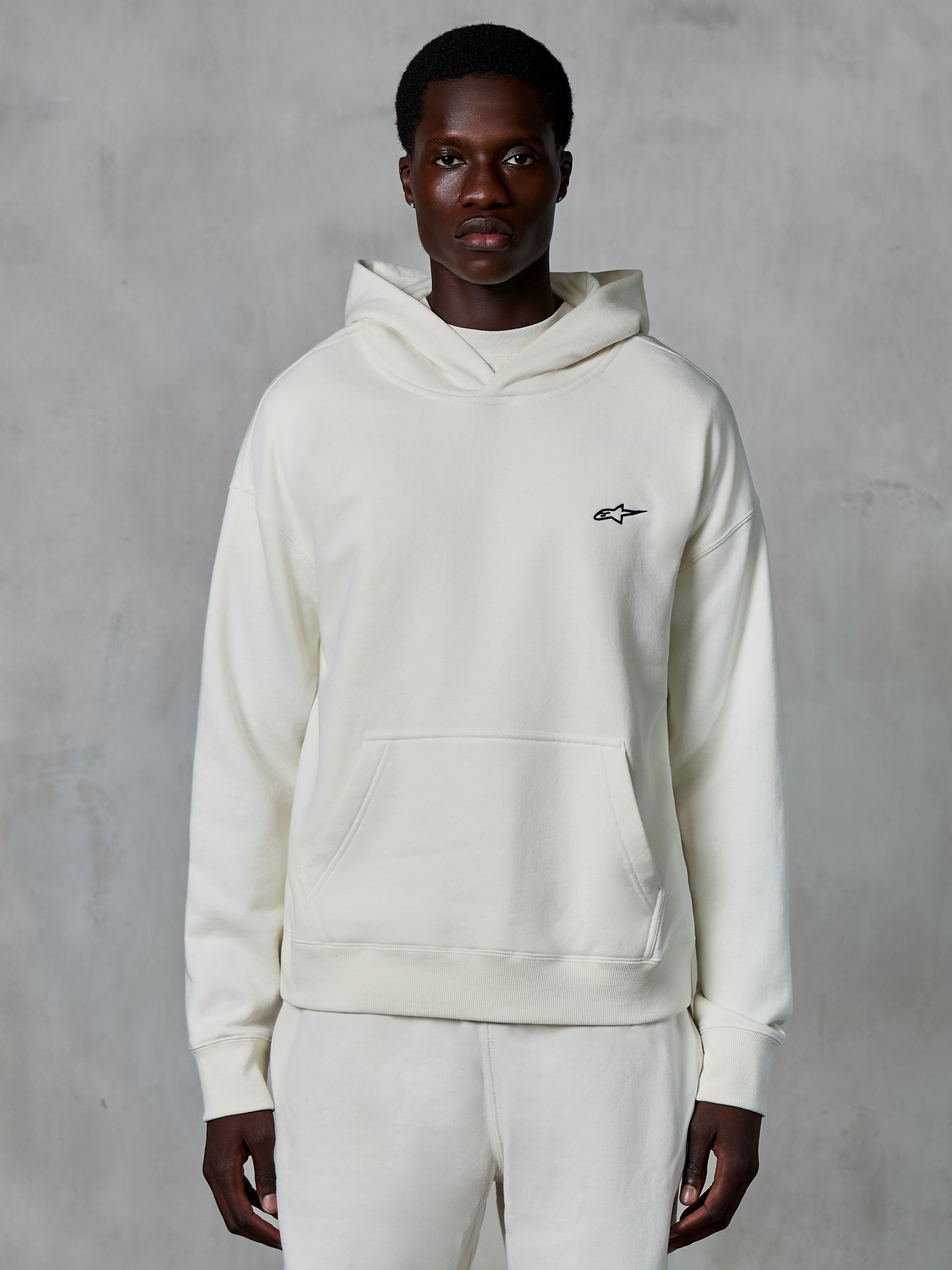 Alpinestars Essentials Ageless Hoodie, Off-White, getragen von einem männlichen Model, kleines schwarzes Logo auf der Brust, Kängurutasche vorne, lange Ärmel, lässige, vom Motorsport inspirierte Streetwear