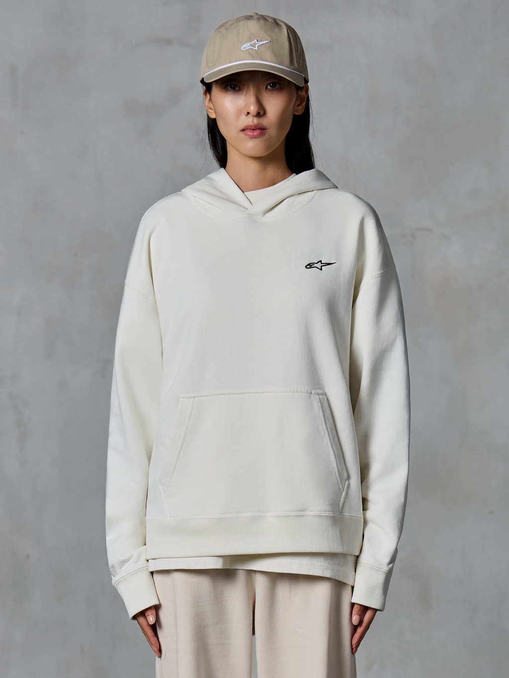 Alpinestars Essentials Ageless Hoodie, cremeweißer Fleece-Kapuzenpullover, weibliches Model mit beiger Kappe und passender cremefarbener Jogginghose, kleines schwarzes Alpinestars-Logo auf der Brust, Kängurutasche auf der Vorderseite, entspannte Passform, lässige Motorsport-inspirierte Streetwear
