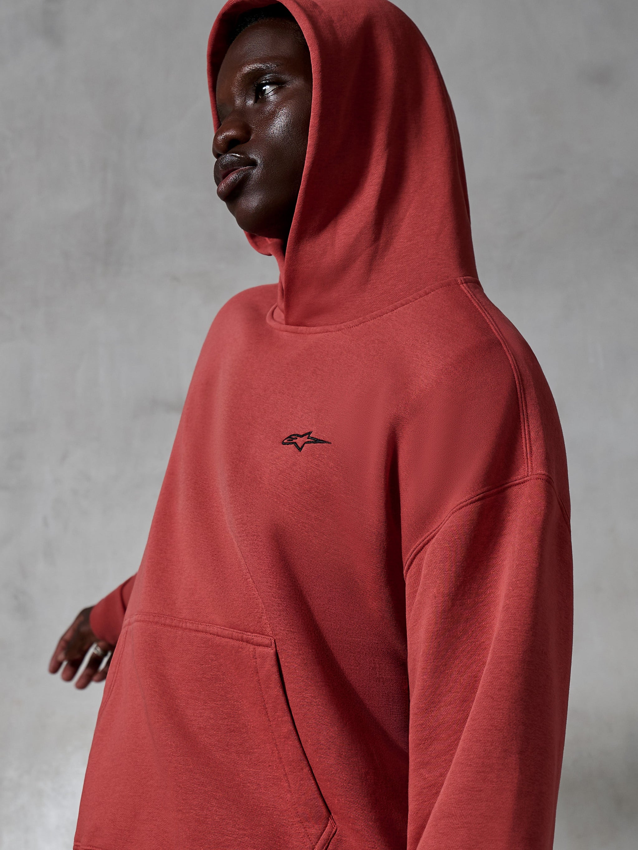 Alpinestars Essentials Ageless Hoodie, casual sportswear, Brick red, worn by a model with hood up, featuring a small black embroidered logo, soft fleece texture, relaxed fit streetwear