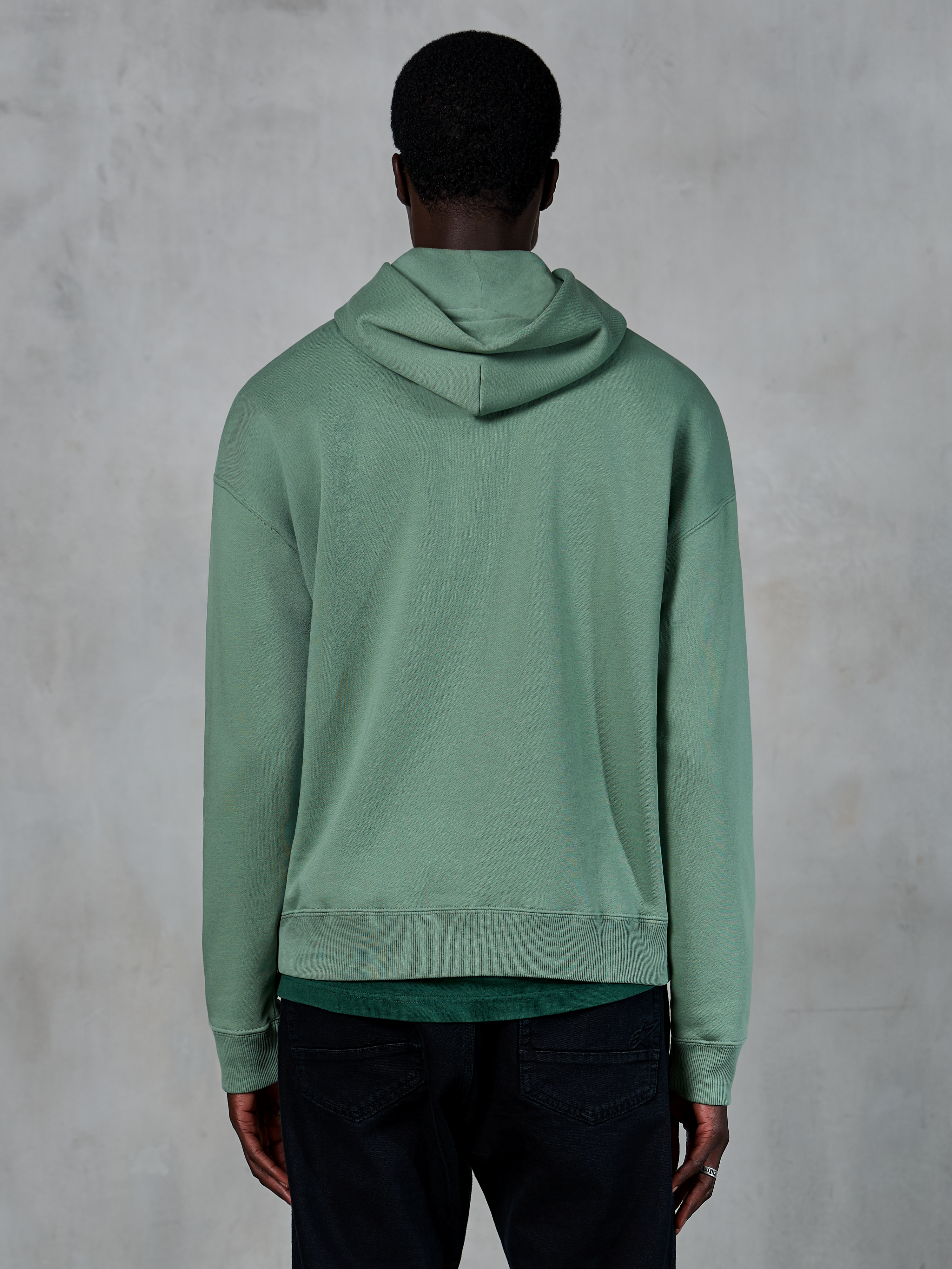 Alpinestars Essentials Ageless Hoodie, lässige Sportbekleidung, Kaktusgrün, Rückansicht am Modell, Garment-Stone-Wash-Finish, kombiniert mit schwarzer Hose, minimalistisches Lifestyle-Design