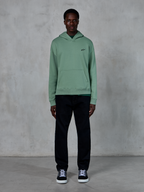 Alpinestars Essentials Ageless Hoodie, Kaktusgrün, kleines schwarzes Brustlogo, Kängurutasche, lässige Lifestyle-Streetwear getragen mit schwarzer Hose und schwarz-weißen Sneakern vor einer strukturierten Wand