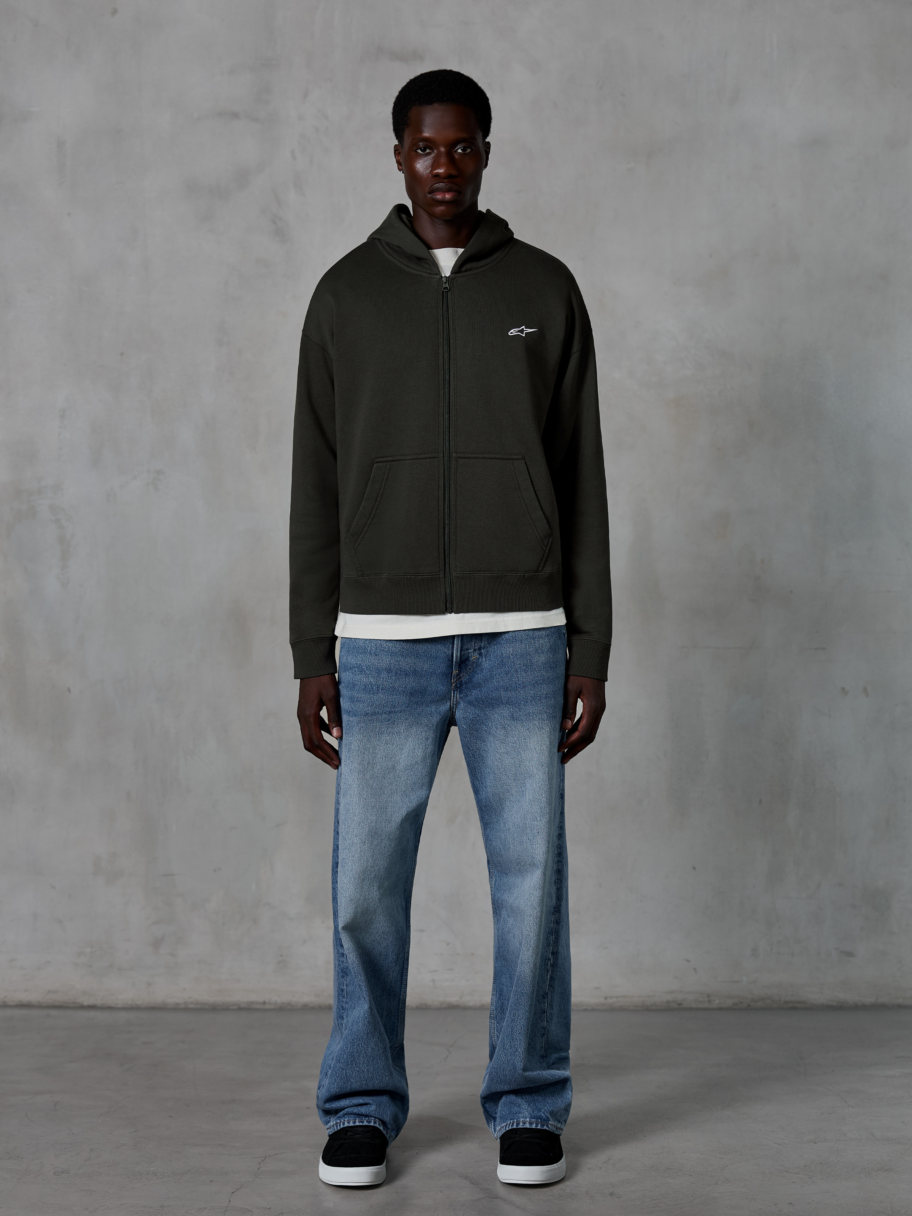Alpinestars Essentials Ageless Zip Hoodie, Kapuzenjacke mit Reißverschluss, Off Black, mit einem kleinen weißen Alpinestars-Logo auf der Brust, getragen von einem Model mit weißem T-Shirt und blauen Jeans, entspannte Passform, lässige Motorsport-Streetwear