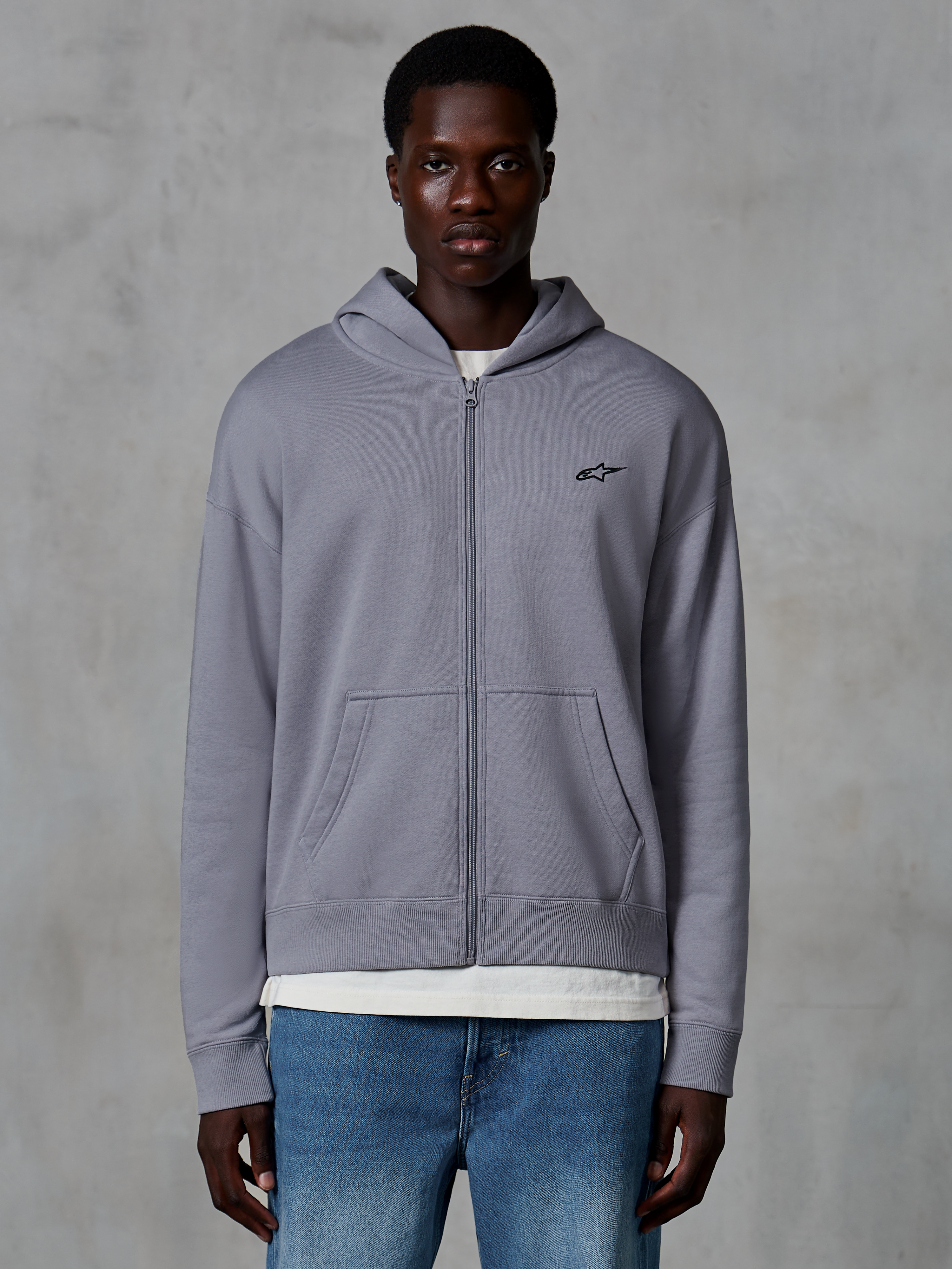 Alpinestars Essentials Ageless Zip Hoodie, Silber, lässige Streetwear von einem Model getragen, Fleece-Jacke mit durchgehendem Reißverschluss und kleinem schwarzen Astars-Logo auf der Brust, Kängurutaschen, entspannte Passform