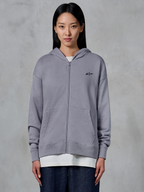 Alpinestars Essentials Ageless Zip Hoodie, Silber, lässige Streetwear, Fleece-Sweatshirt mit durchgehendem Reißverschluss und kleinem schwarzem Alpinestars-Logo auf der Brust, geteilte Kängurutasche, von einem Model über einem weißen T-Shirt und dunklen Jeans getragen