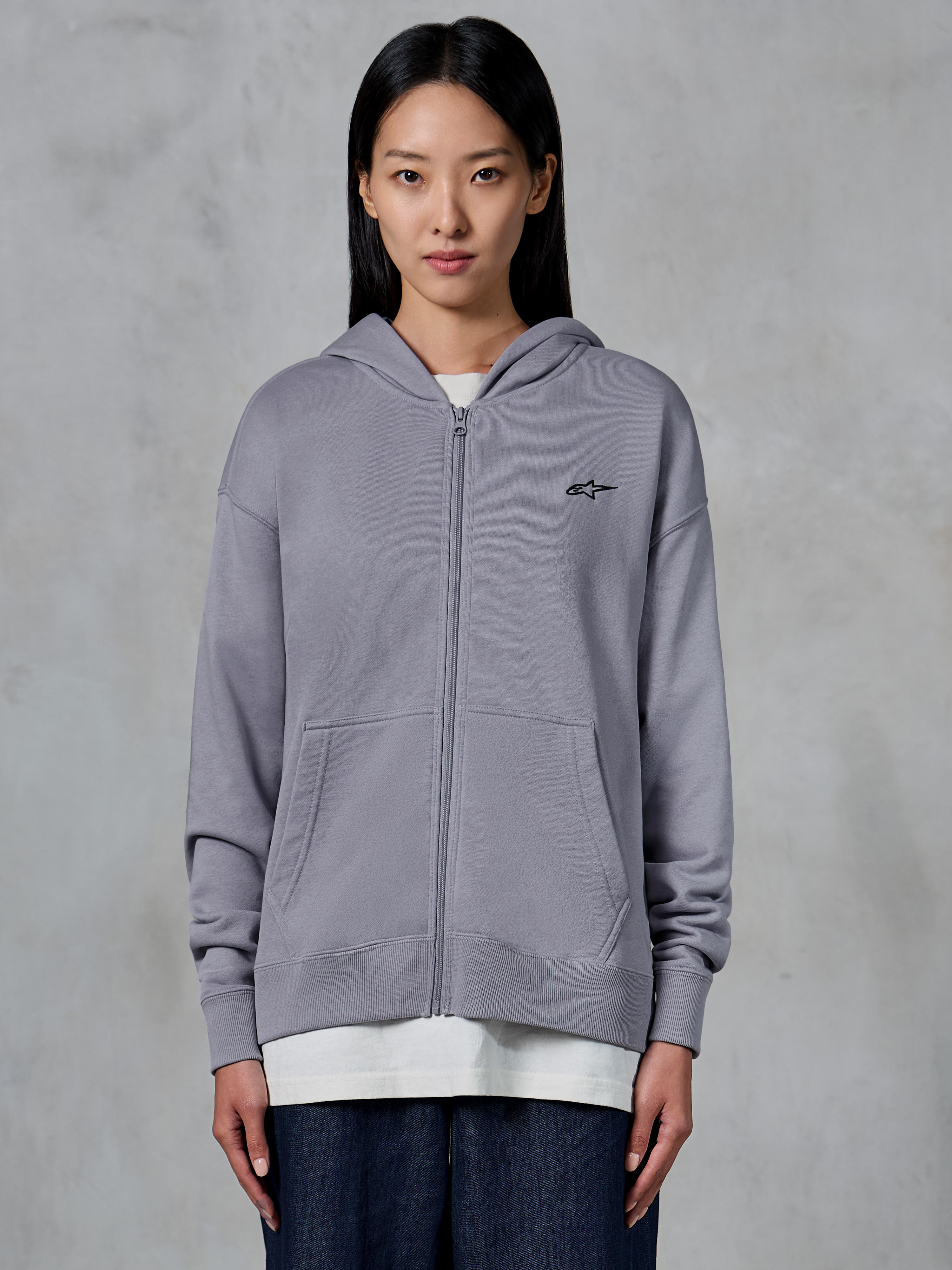 Alpinestars Essentials Ageless Zip Hoodie, Silber, lässige Streetwear, Fleece-Sweatshirt mit durchgehendem Reißverschluss und kleinem schwarzem Alpinestars-Logo auf der Brust, geteilte Kängurutasche, von einem Model über einem weißen T-Shirt und dunklen Jeans getragen