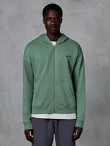 Alpinestars Essentials Ageless Zip Hoodie, casual streetwear zip-up, Cactus green, featuring a subtle embroidered chest logo, split kangaroo pockets, and ribbed cuffs, worn by a male model against a textured grey background