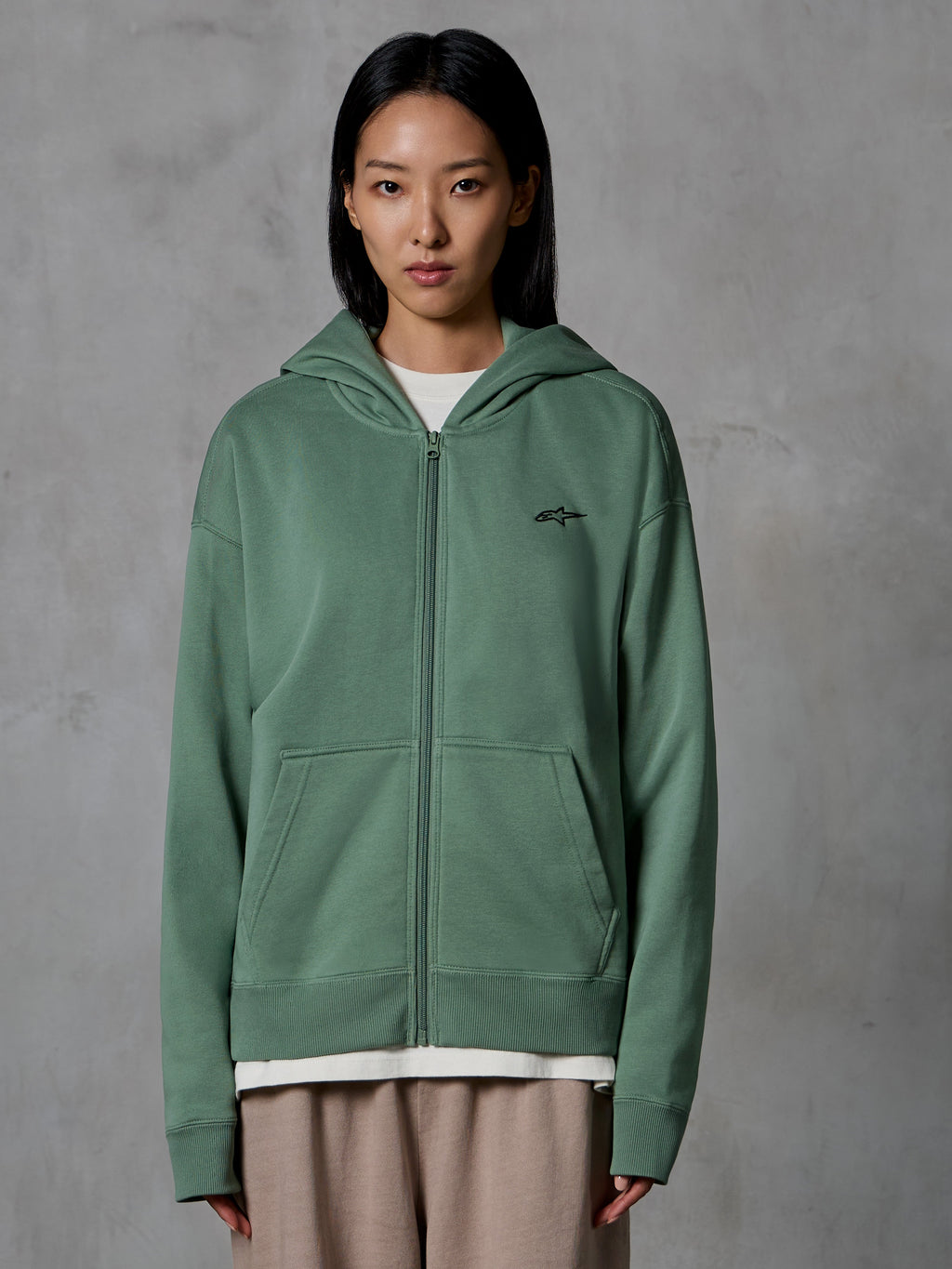 Alpinestars Essentials Ageless Zip Hoodie, Kaktusgrün, legere Streetwear getragen von einem weiblichen Model, Fleece-Sweatshirt mit Reißverschluss und kleinem schwarzen Logo auf der Brust, Fronttaschen, entspannte Passform für den Alltag