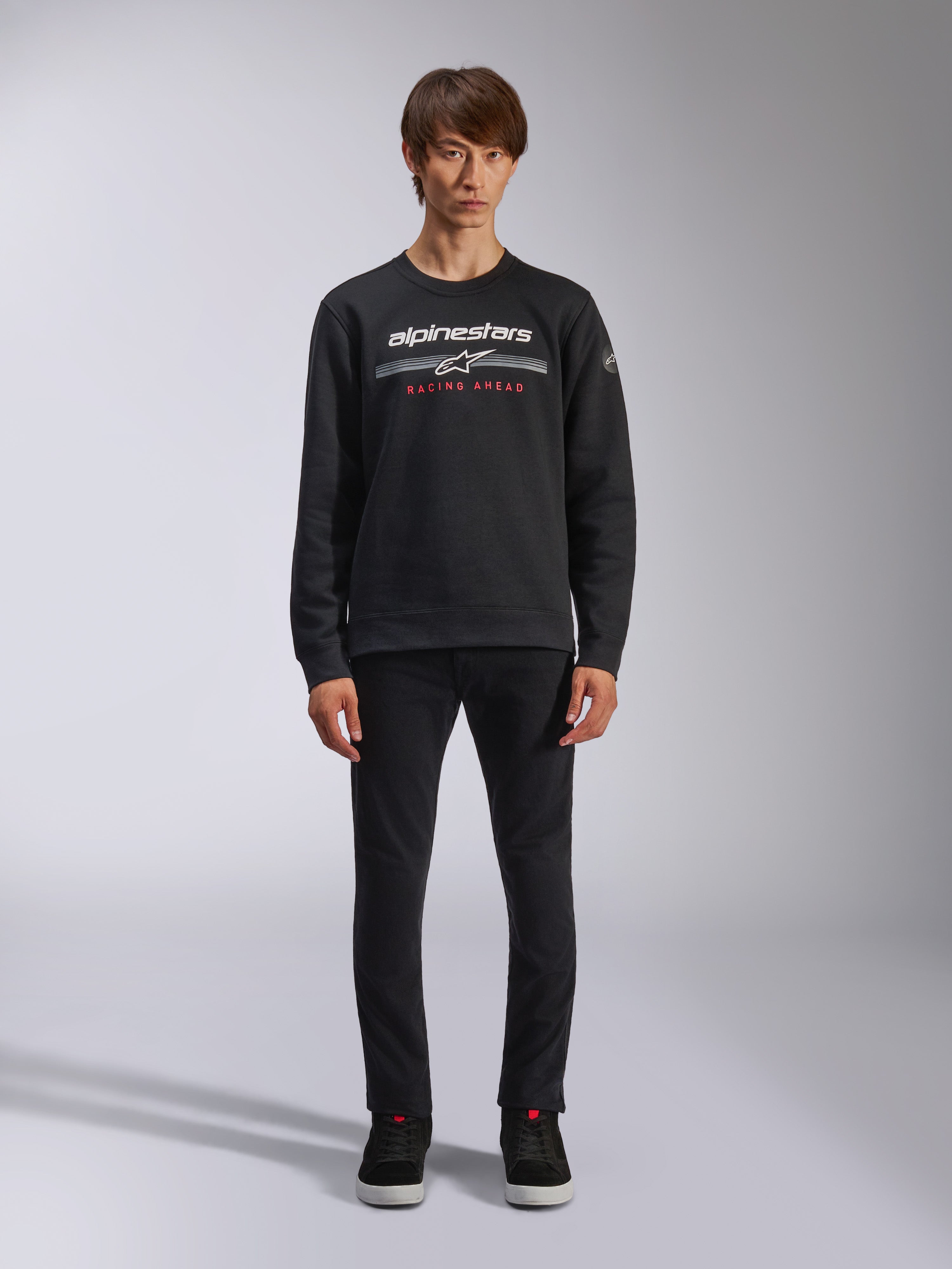 Alpinestars Bettern Crew, schwarzes Sweatshirt mit Rundhalsausschnitt, zeigt weißes Alpinestars-Logo mit grauen horizontalen Streifen und rotem „RACING AHEAD“-Text, lässige Motorsport-Streetwear, getragen von einem Model mit schwarzer Hose und Sneakern
