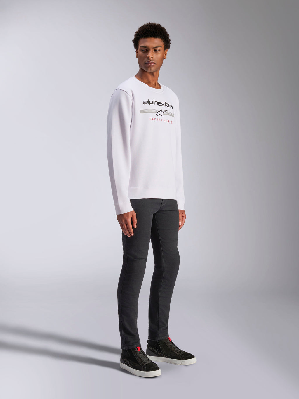 Alpinestars Bettern Crew, weißes Pullover-Sweatshirt mit schwarzem Alpinestars-Logo und rotem „Racing Ahead“-Schriftzug, lässige Motorsport-Streetwear, getragen von einem Model mit dunkler Hose und schwarzen Sneakern