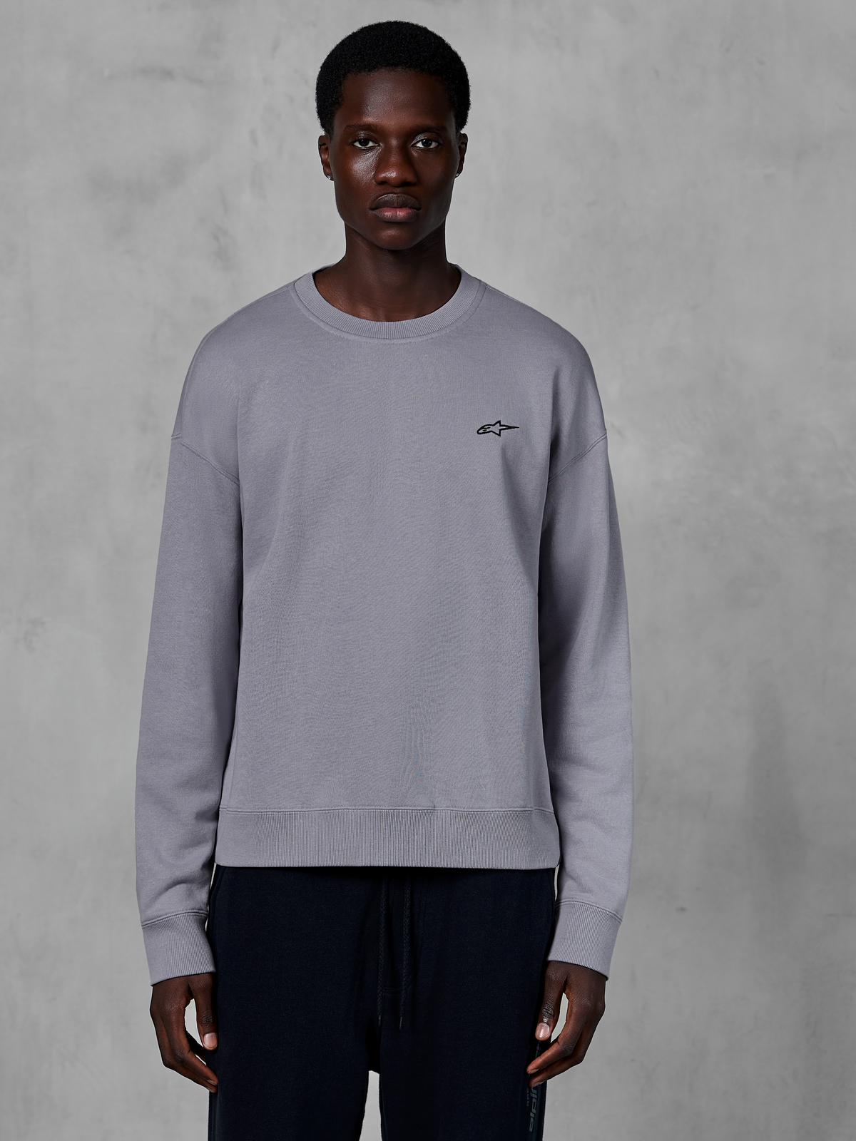 Alpinestars Essentials Ageless Crew, silbernes Rundhals-Sweatshirt von einem Model getragen, mit einem kleinen schwarzen Stern-Logo auf der Brust, entspannte Passform mit Rippbündchen und Saum, lässige Motorsport-Streetwear