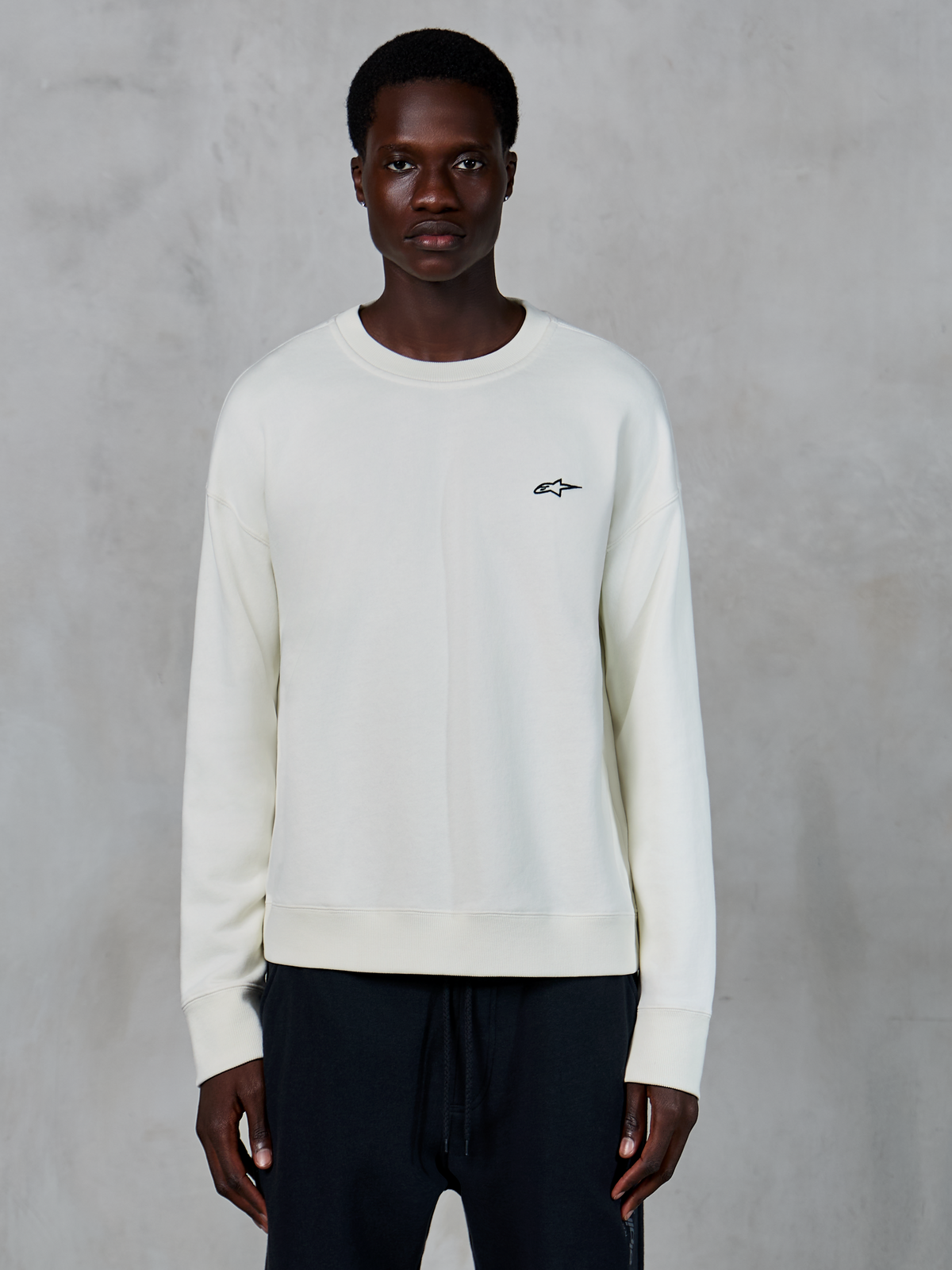 Alpinestars Essentials Ageless Crew, Off-White Rundhals-Sweatshirt, kleines schwarzes Astars-Logo auf der linken Brust, lässige Lifestyle-Sportbekleidung, hochwertige Fleece-Textur, getragen von Model mit schwarzen Jogginghosen