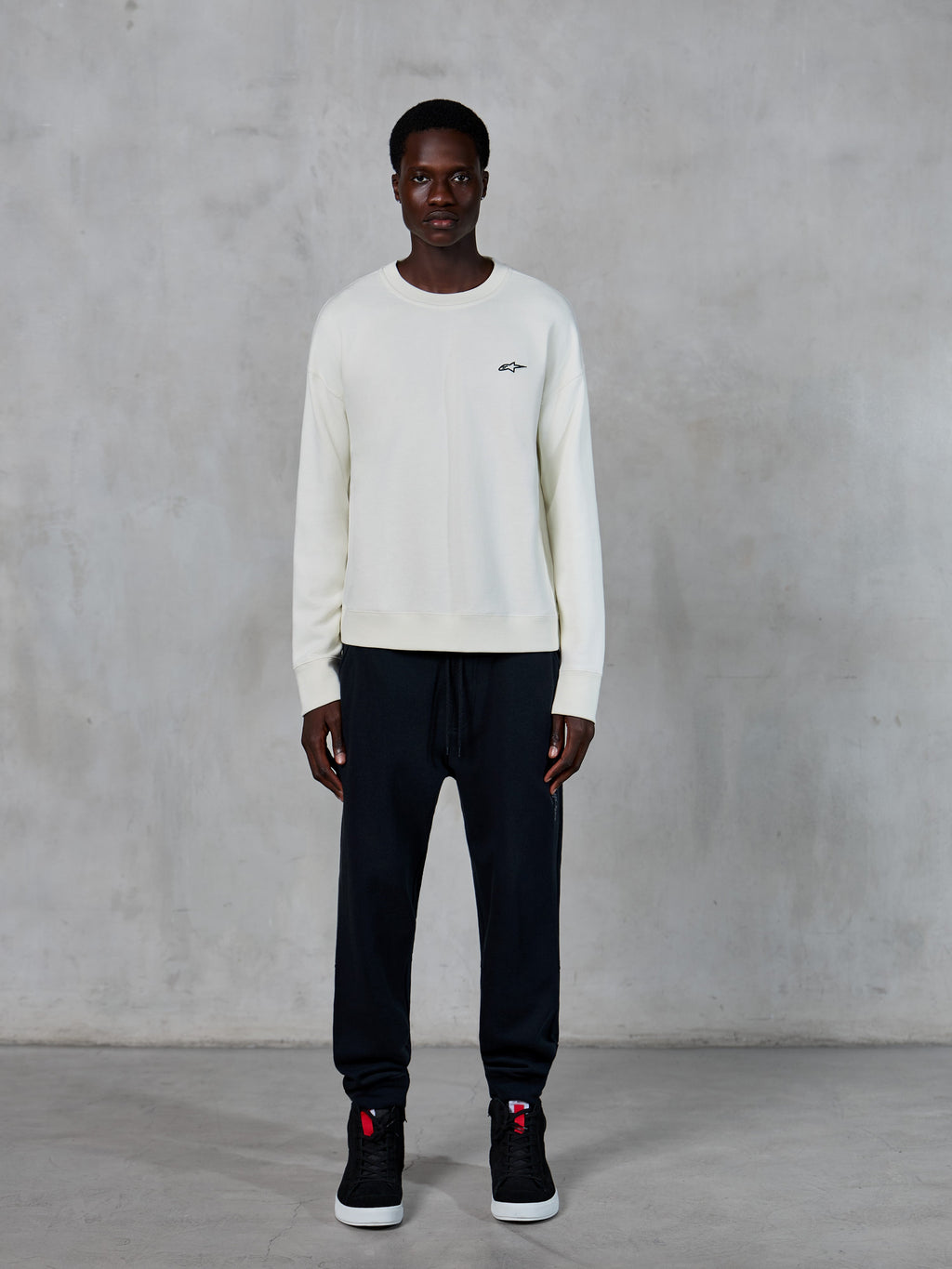 Alpinestars Essentials Ageless Crew, Rundhals-Sweatshirt, Off-White mit kleinem schwarzem Stern-Logo, getragen von Model mit schwarzer Jogginghose, lässige, vom Motorsport inspirierte Streetwear