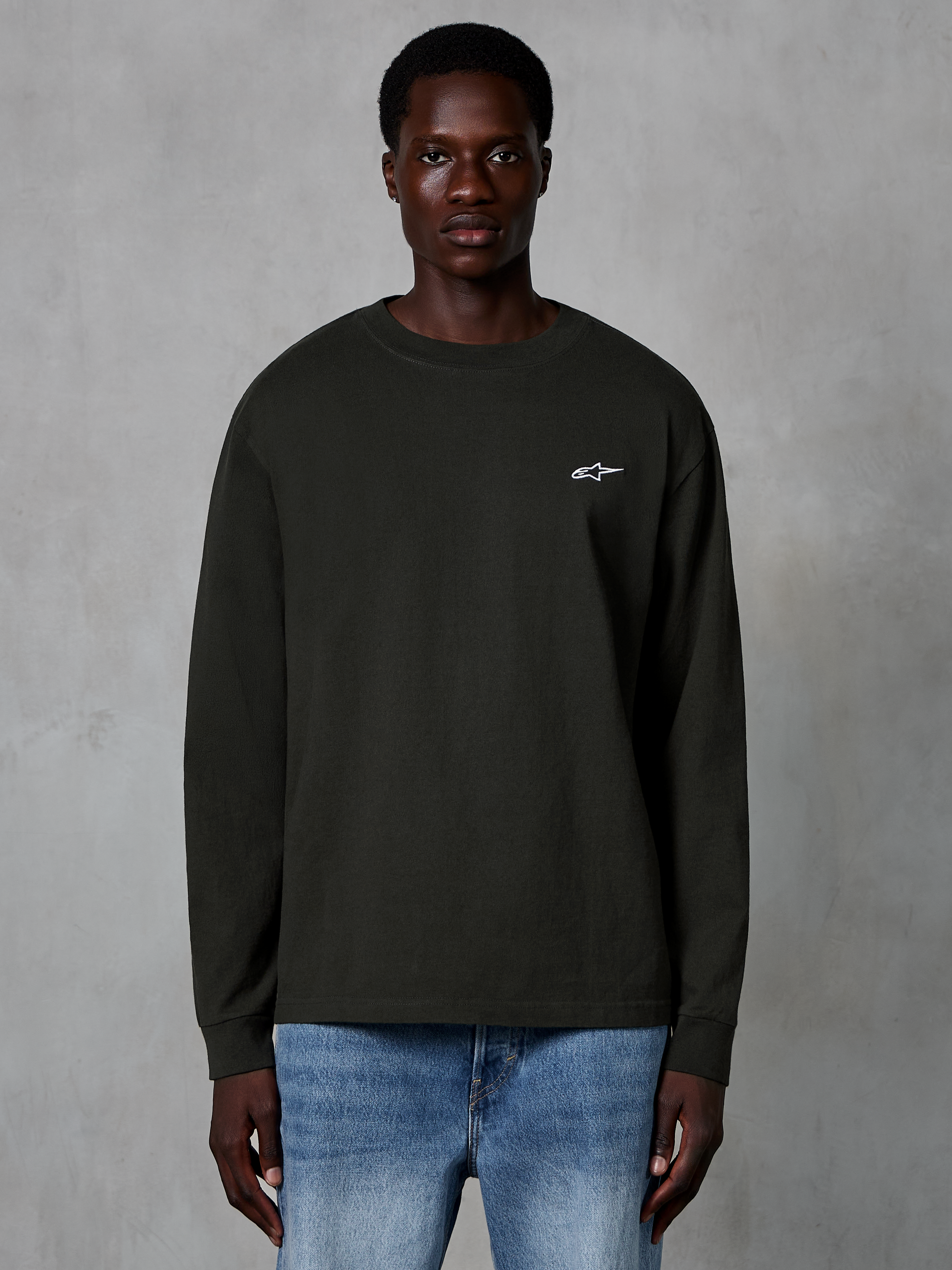 Alpinestars Essentials Ageless LS Tee, Langarm-T-Shirt, Off-Black, mit kleinem weißen Alpinestars-Logo auf der Brust, getragen von einem Modell mit blauen Jeans, lässige Streetwear aus Bio-Baumwolle