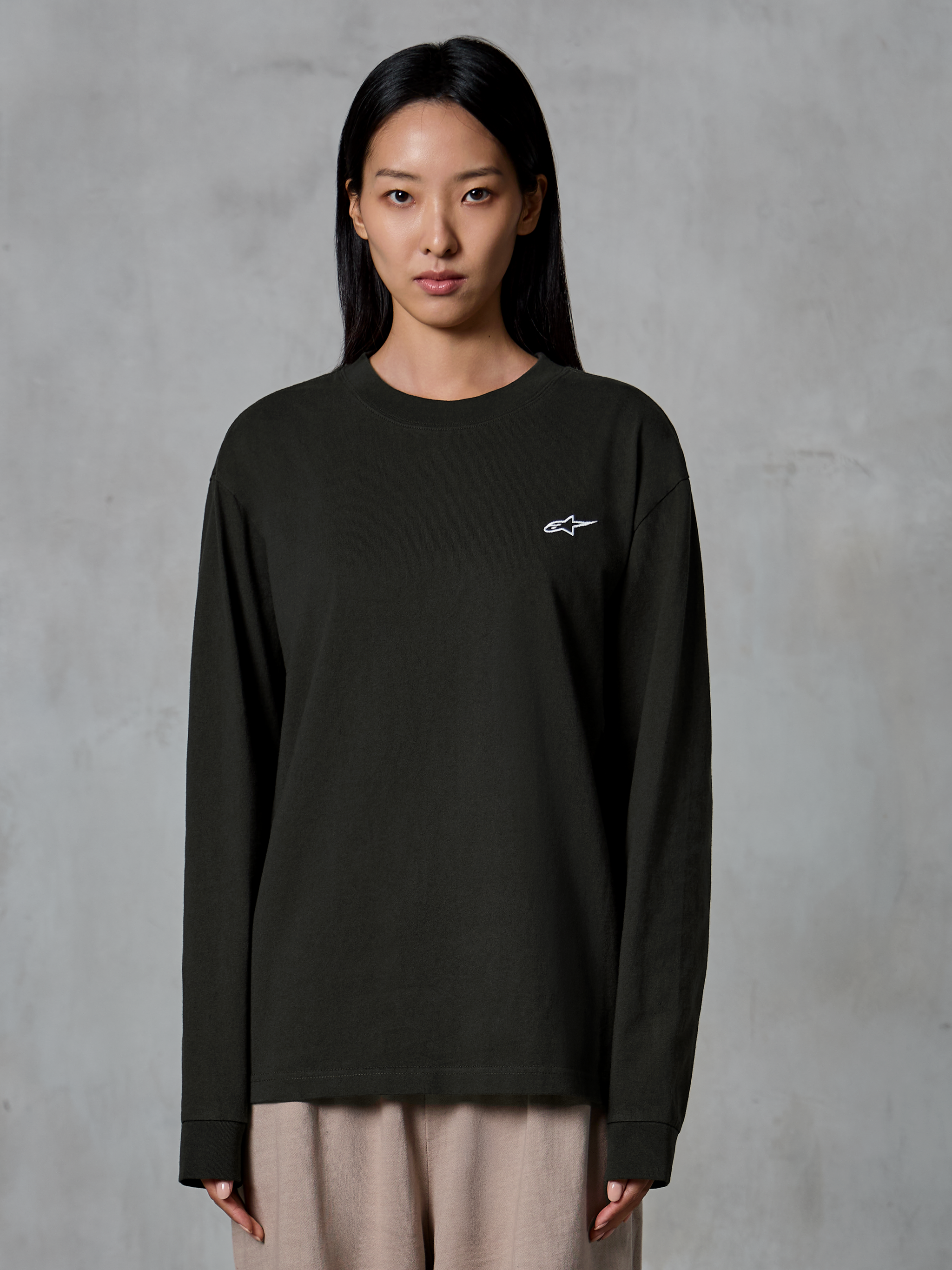 Model im Alpinestars Essentials Ageless LS Tee, Langarmshirt, Off-Black, mit einem kleinen weißen Alpinestars-Logo auf der Brust, kombiniert mit einer beigen Hose, lässige Lifestyle-Streetwear