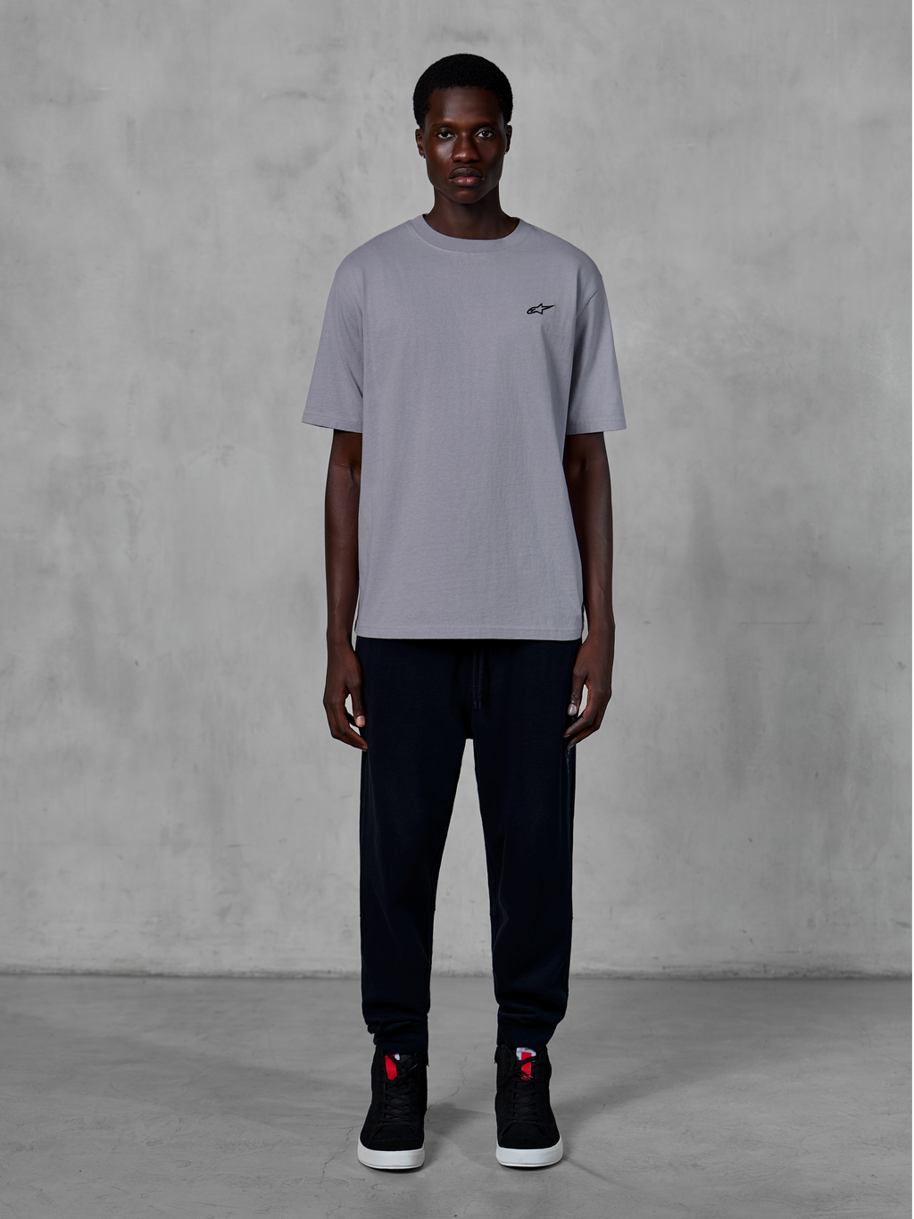Alpinestars Essentials Ageless SS Tee, Kurzarm-T-Shirt, Silber, mit einem kleinen schwarzen Logo auf der Brust, kombiniert mit schwarzen Jogginghosen und Sneakern, lässige Motorsport-Streetwear