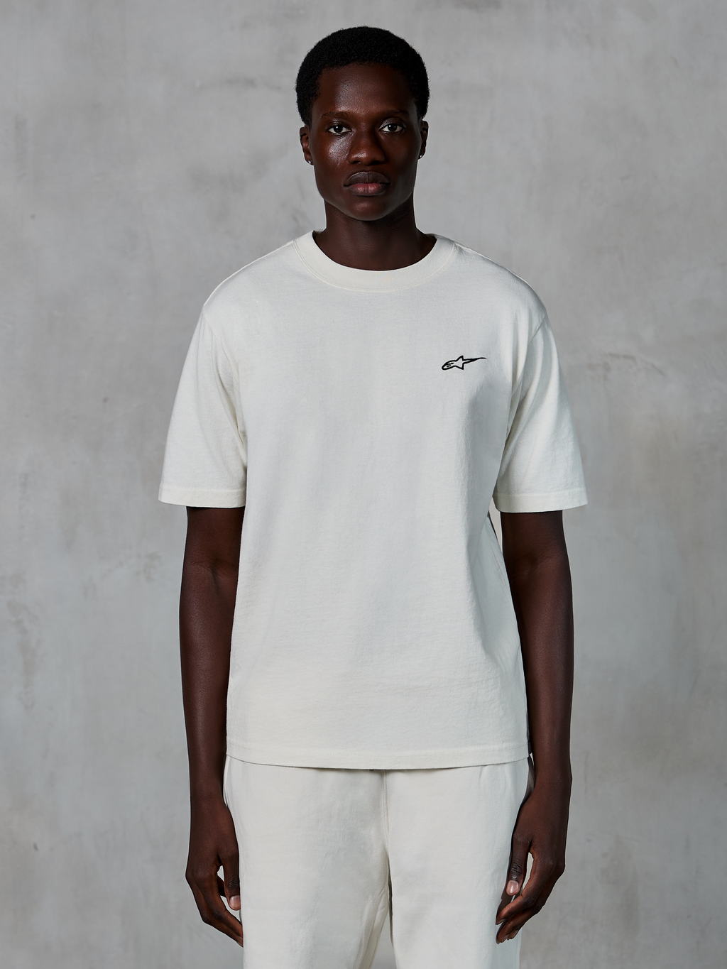 Alpinestars Essentials Ageless SS Tee, Kurzarm-T-Shirt, Off-White, von einem männlichen Model getragen, mit einem kleinen schwarzen Alpinestars-Logo auf der Brust, entspannte Passform, Bio-Baumwolljersey, lässige Motorsport-Streetwear