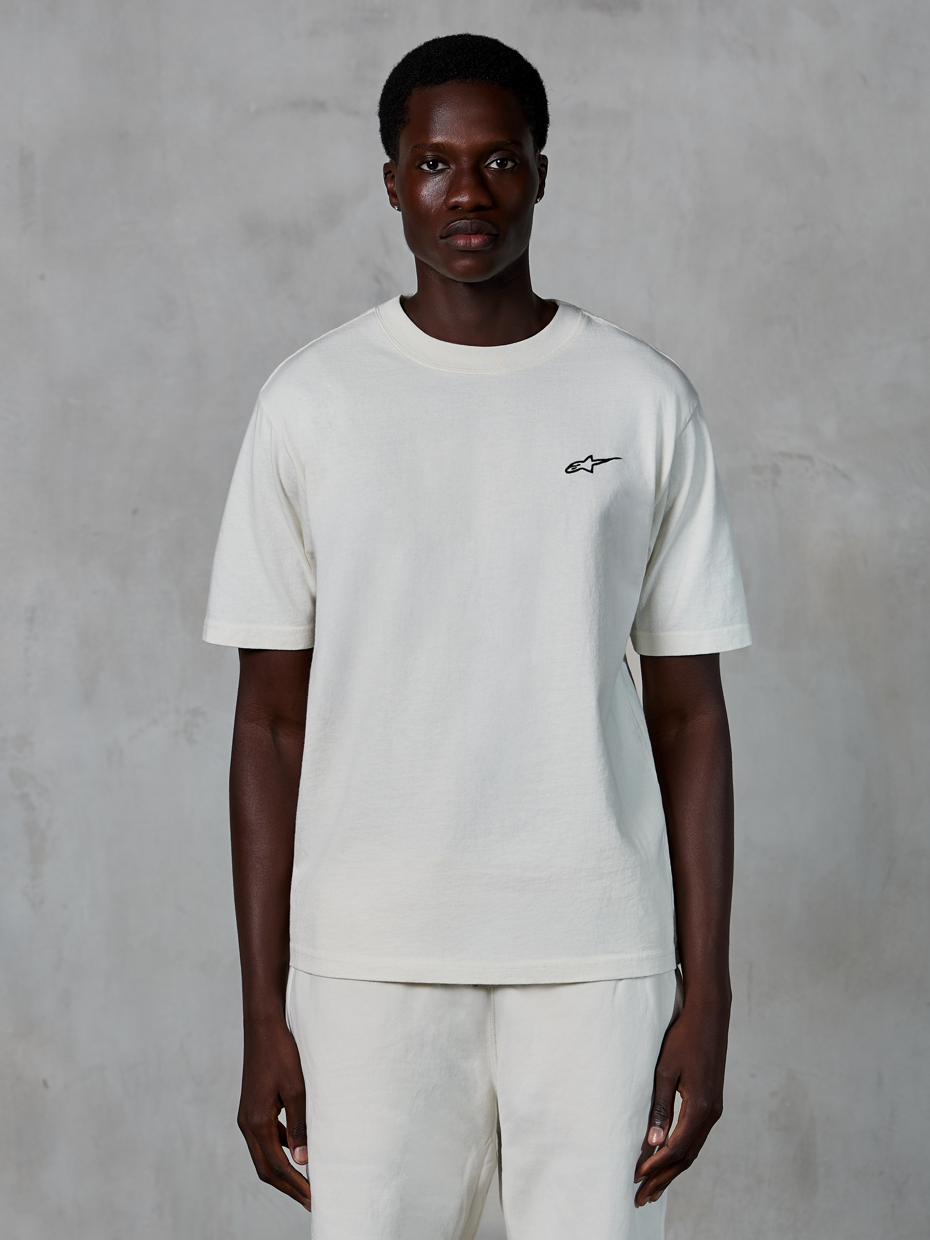 Alpinestars Essentials Ageless SS Tee, Kurzarm-T-Shirt, Off-White, von einem männlichen Model getragen, mit einem kleinen schwarzen Alpinestars-Logo auf der Brust, entspannte Passform, Bio-Baumwolljersey, lässige Motorsport-Streetwear