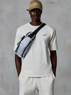Alpinestars Essentials Ageless SS Tee, Kurzarm-T-Shirt, Off-White, getragen mit passenden Shorts und einer hellbraunen Kappe, mit einem kleinen schwarzen Logo auf der Brust und einer grauen Umhängetasche, lässige, vom Motorsport inspirierte Streetwear