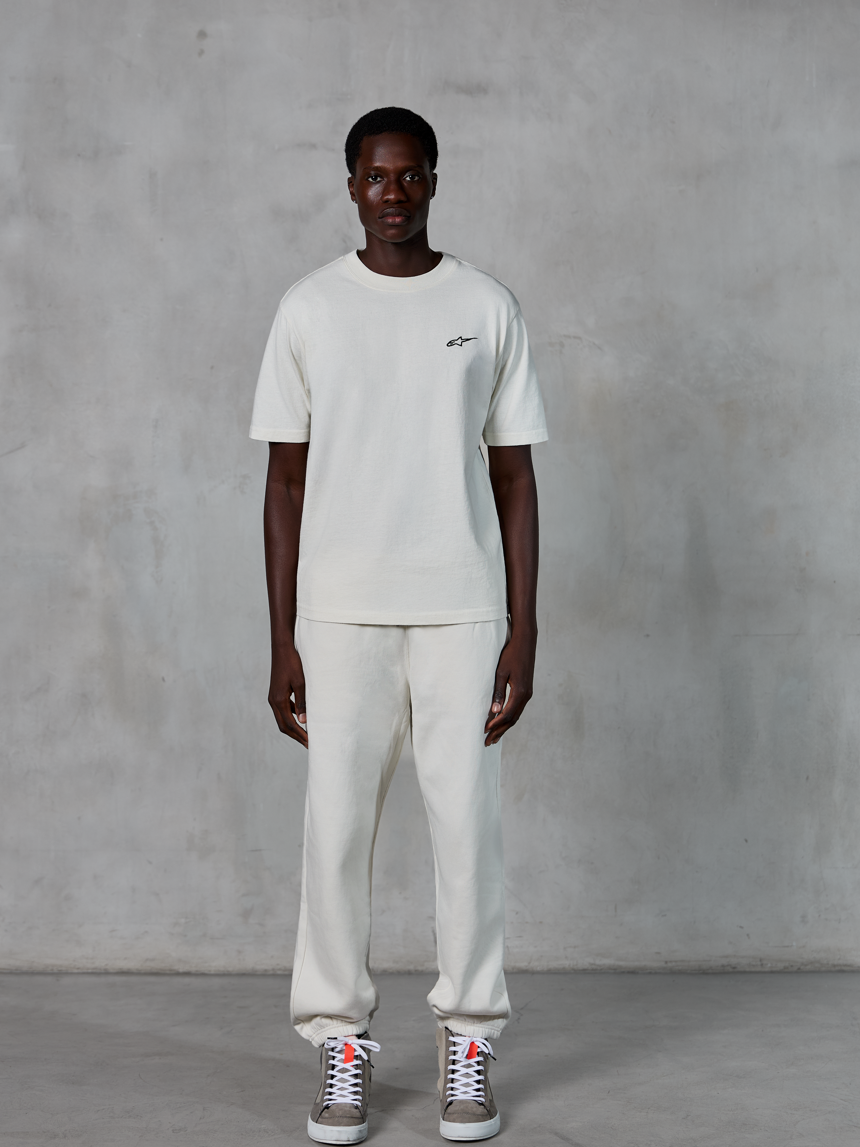 Alpinestars Essentials Ageless SS Tee, kurzärmeliges T-Shirt, Off-White, mit kleinem schwarzen Alpinestars-Logo auf der Brust, kombiniert mit passender Jogginghose und Sneakern für einen entspannten Lifestyle-Look