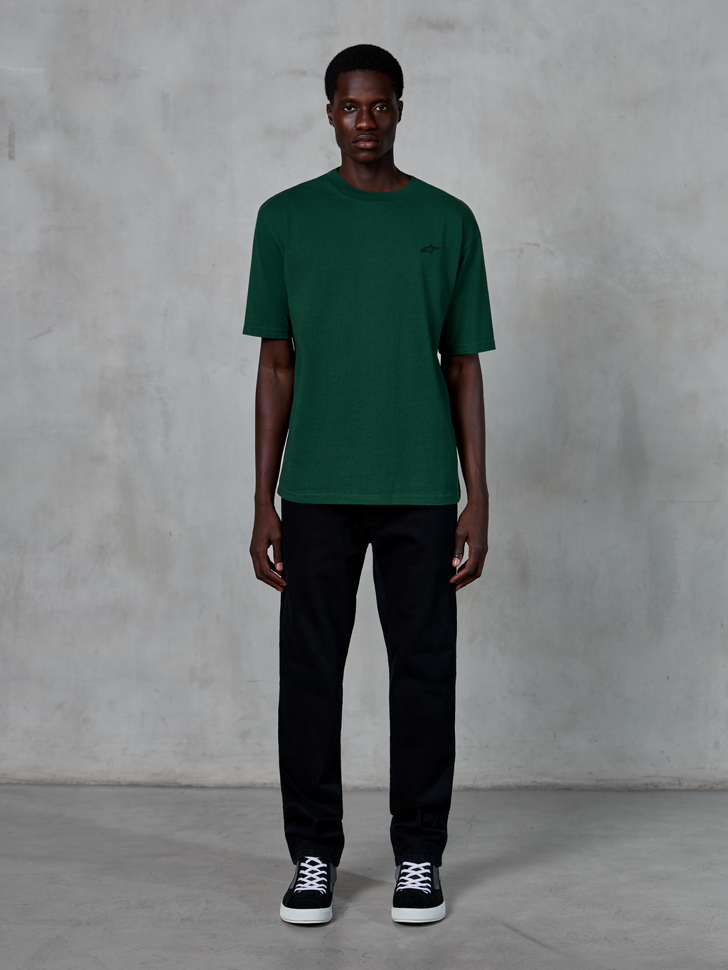 Alpinestars Essentials Ageless SS Tee, Kurzarm-T-Shirt, Pine Green, mit dezentem schwarzem Brustlogo, getragen von einem Model mit schwarzer Hose und Sneakern, lässige Lifestyle-Streetwear mit entspannter Passform