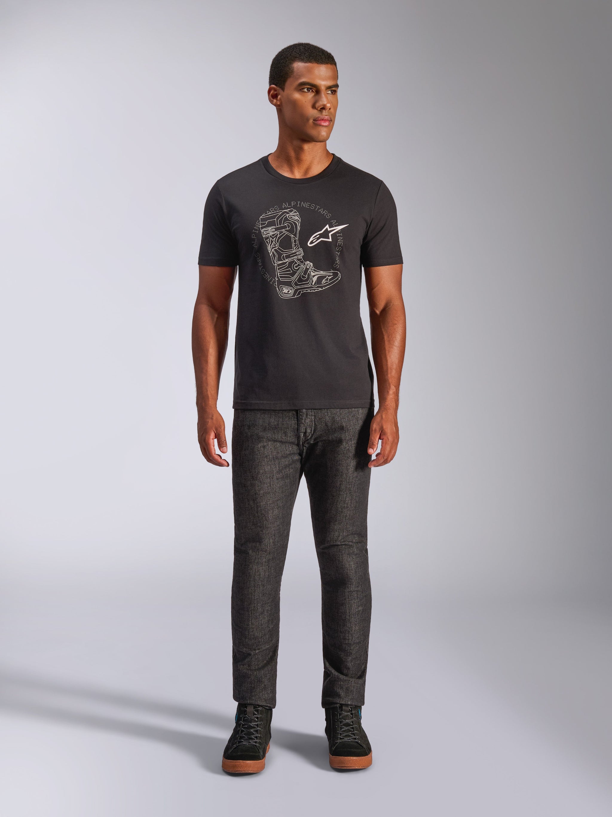 Alpinestars Tech Boot CSF Tee, Short Sleeve T-Shirt, Black, featuring a white graphic of a motocross boot and logo on the chest, worn by a model with dark grey trousers and black sneakers, casual motorsport-inspired design