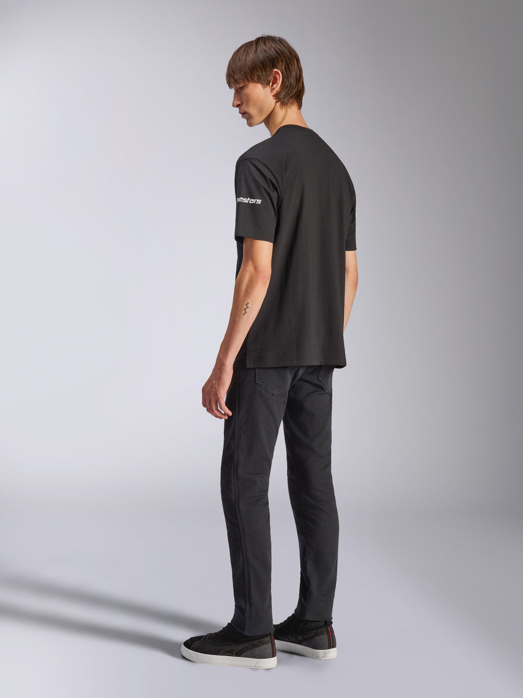 Alpinestars Frontal CSF T-Shirt, kurzärmeliges T-Shirt, Schwarz, weißes Astars-Branding auf dem Ärmel, getragen von einem Model mit schwarzer Hose und Schuhen, lässiges, vom Motorsport inspiriertes Design