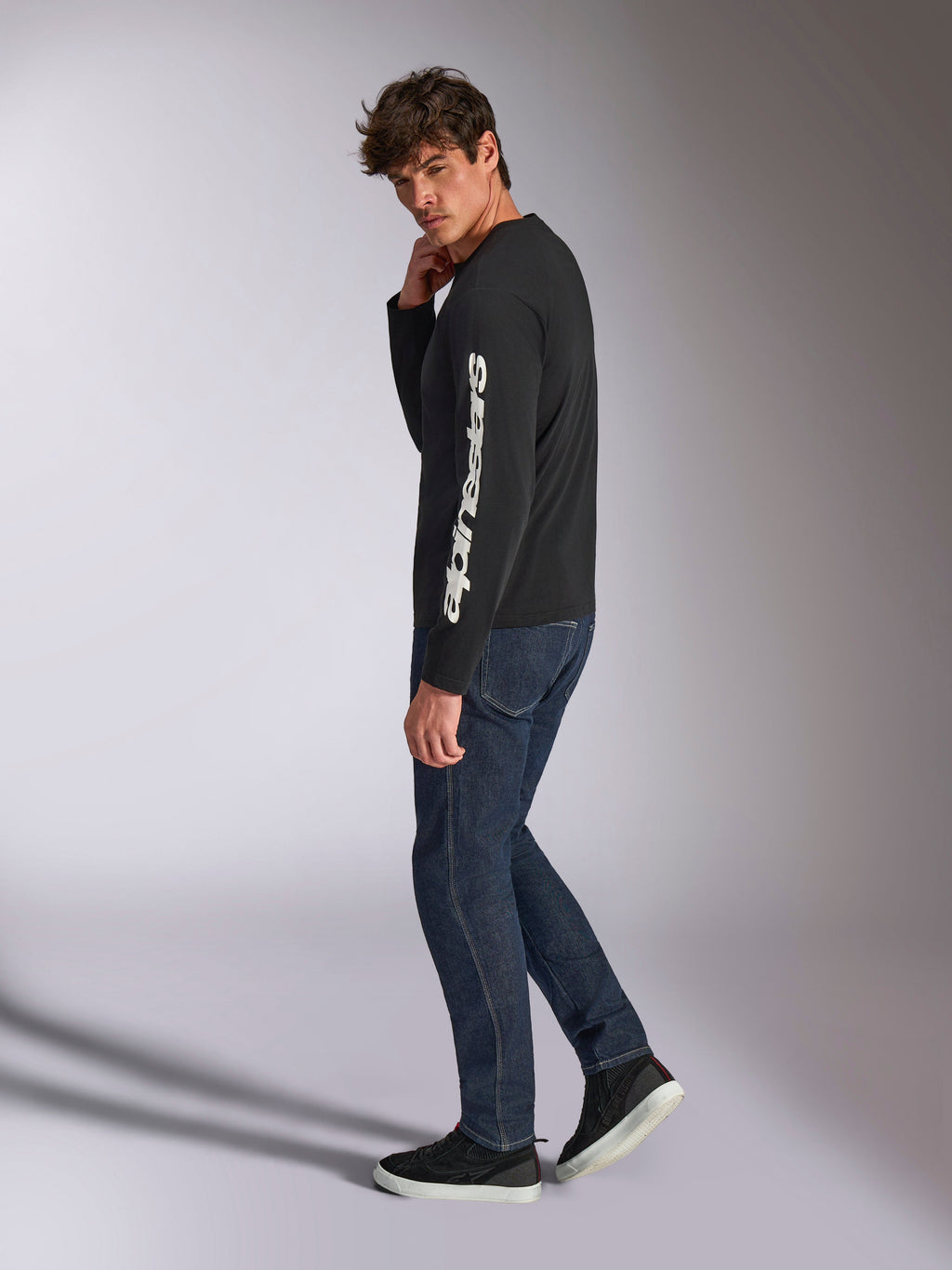 Alpinestars Trackside CSF Langarmshirt, Schwarz mit weißem vertikalem Alpinestars-Schriftzug auf dem Ärmel, getragen von einem Model mit dunkler Jeans und Sneakern, lässige, vom Motorsport inspirierte Sportbekleidung