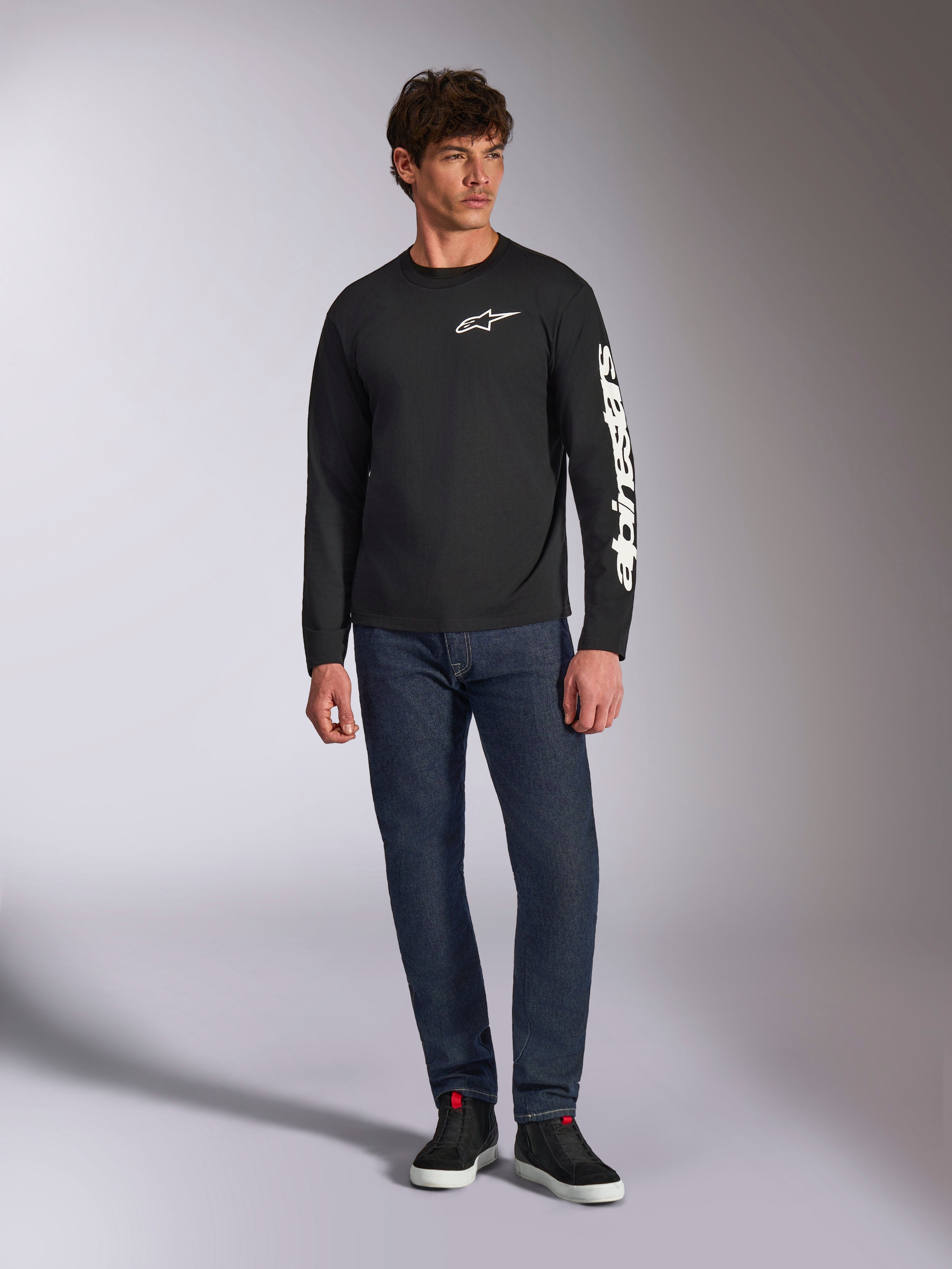Alpinestars Trackside CSF Tee, Langarmshirt, Schwarz und Weiß, getragen mit dunkelblauer Jeans und schwarzen Sneakern, mit weißem Ride 4.0 Logo auf der Brust und Alpinestars-Schriftzug auf dem Ärmel, lässige vom Motorsport inspirierte Streetwear