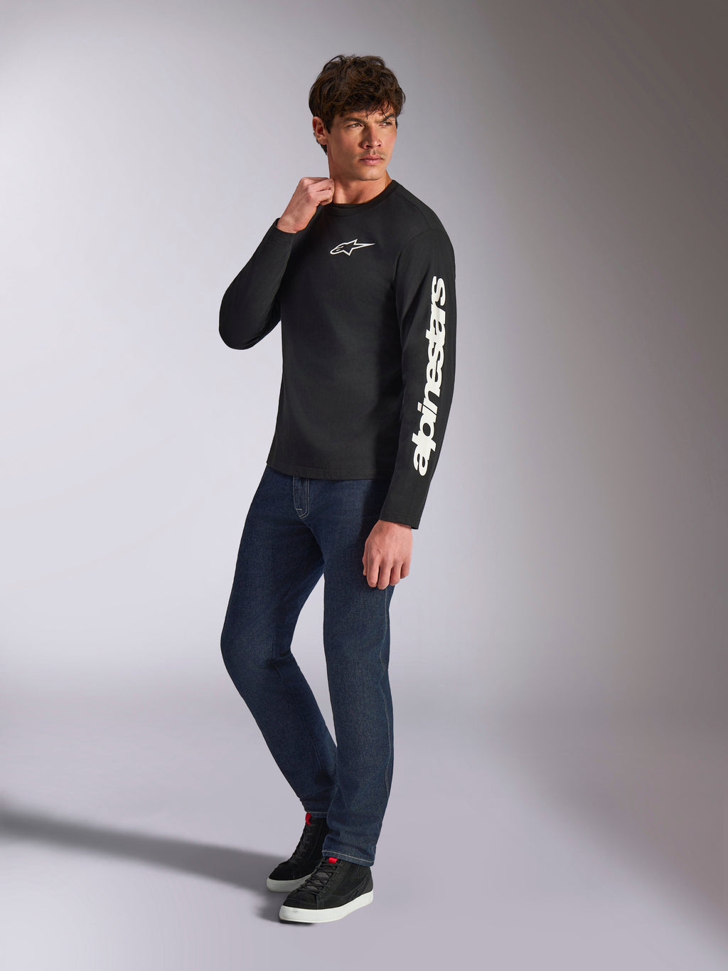 Alpinestars Trackside CSF Tee, Langarmshirt, Schwarz, weißes vertikales Alpinestars-Branding auf dem Ärmel und Brust-Logo, getragen von einem männlichen Model mit dunkelblauen Jeans und Sneakern, lässige Motorsport-Streetwear