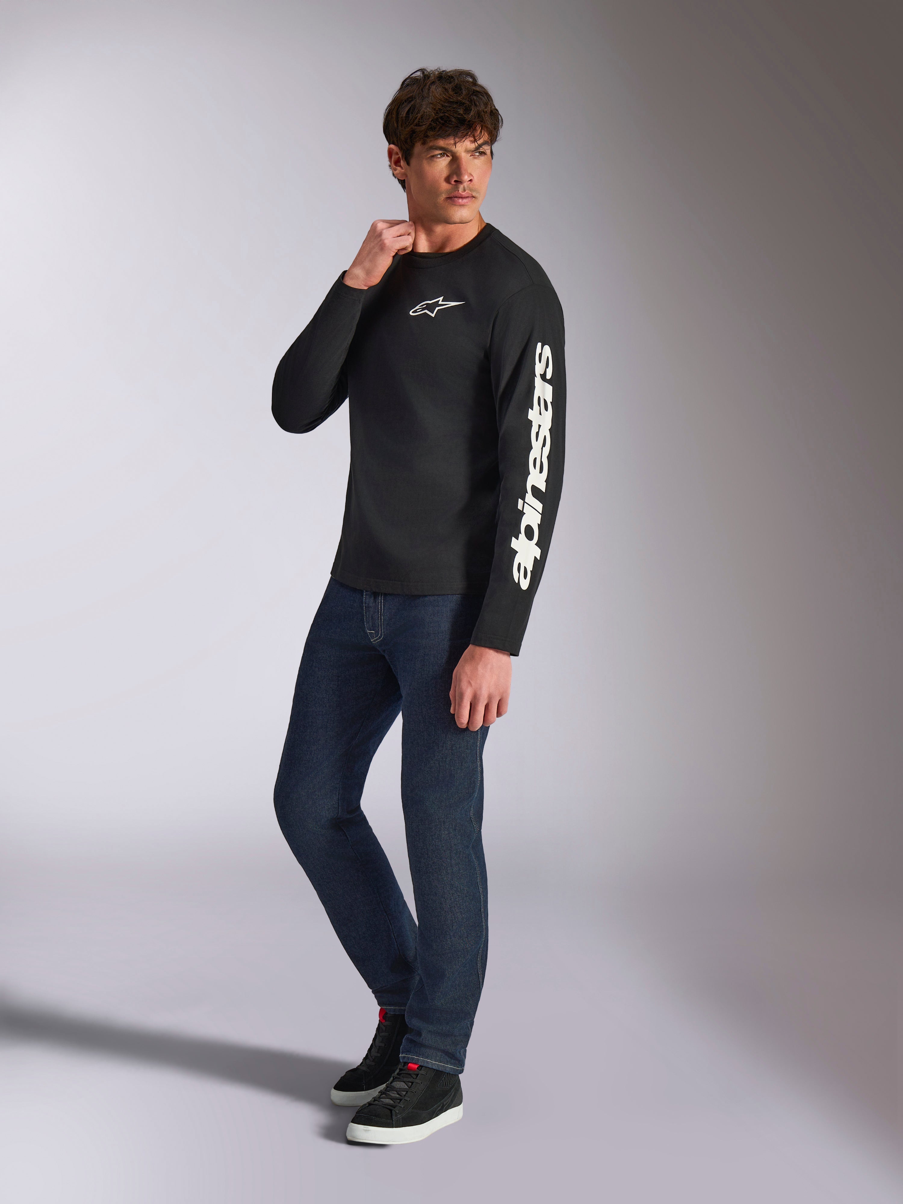 Alpinestars Trackside CSF Tee, Langarmshirt, Schwarz, weißes vertikales Alpinestars-Branding auf dem Ärmel und Brust-Logo, getragen von einem männlichen Model mit dunkelblauen Jeans und Sneakern, lässige Motorsport-Streetwear
