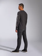 Alpinestars Trackside CSF Langarm-T-Shirt, Schwarz, mit großem grauem Alpinestars-Logo auf dem linken Ärmel, getragen von einem Modell mit schwarzer Hose und lässigen Sneakern, Motorsport-inspirierte Streetwear.
